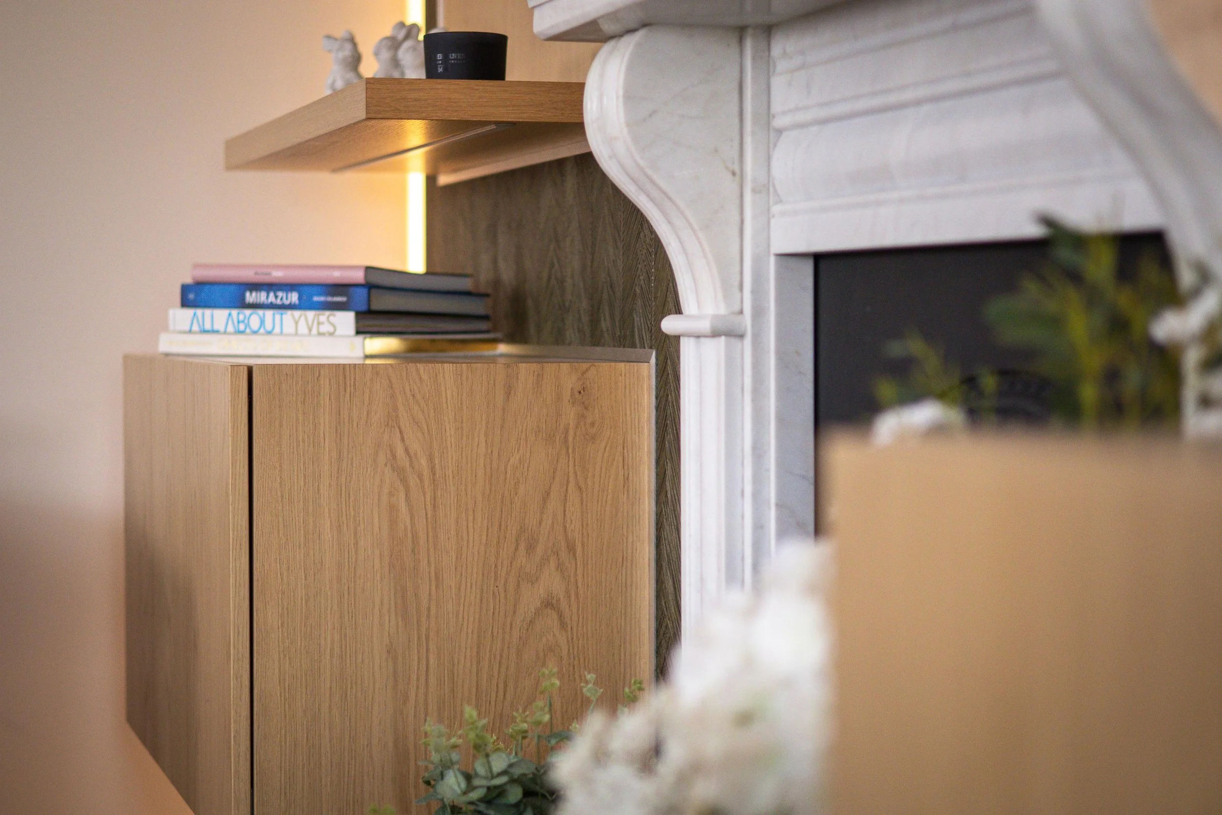 Close-up of a wooden cabinet with decorative rabbits on top, a black container, and a stack of books; part of a fireplace with white mantel and a fireplace opening in the right foreground, and a soft beige wall in the background.