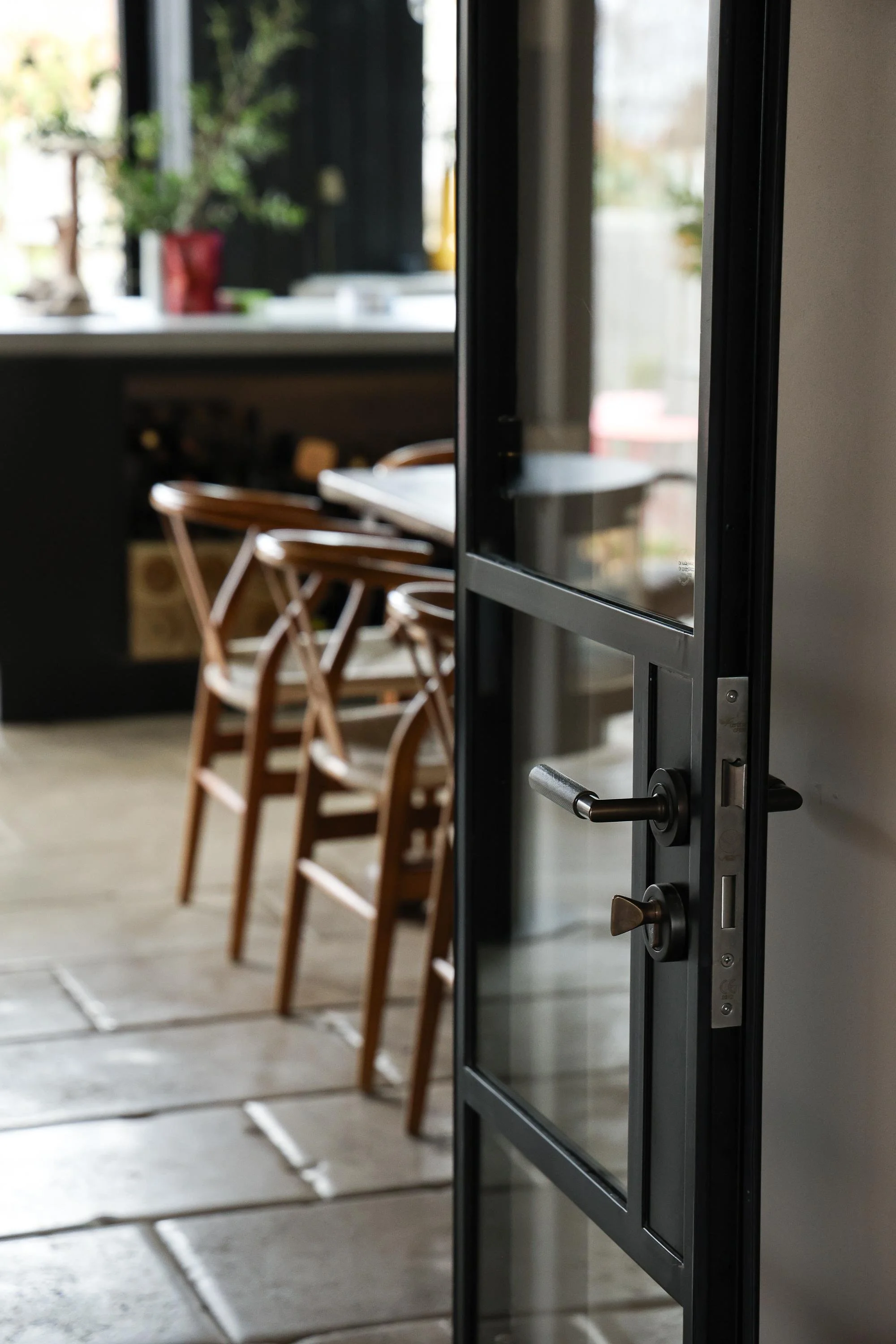 Open glass door leading into a room with wooden chairs, a counter, and a window with plants outside.