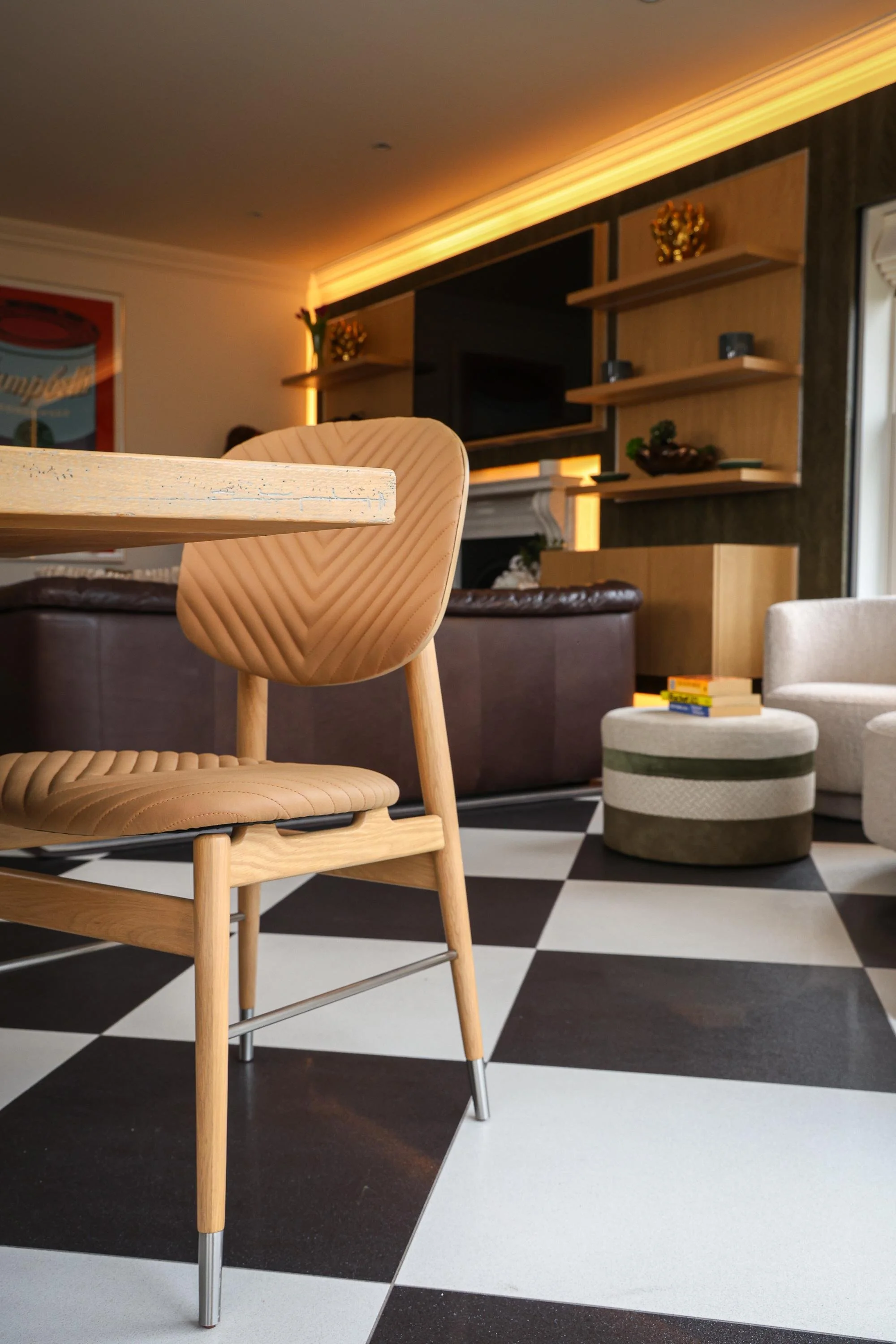 A modern interior with a wooden chair in the foreground, black and white checkered flooring, and a seating area with a round ottoman and a white armchair.