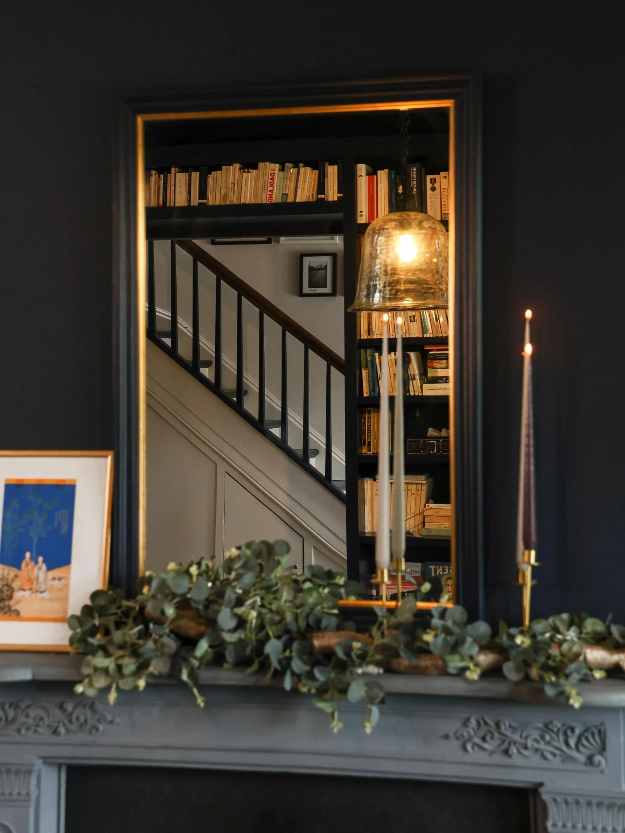 A mirror reflects a room with a bookshelf filled with books, a framed black and white photo on the wall, a staircase with a dark railing, and a lit pendant light.