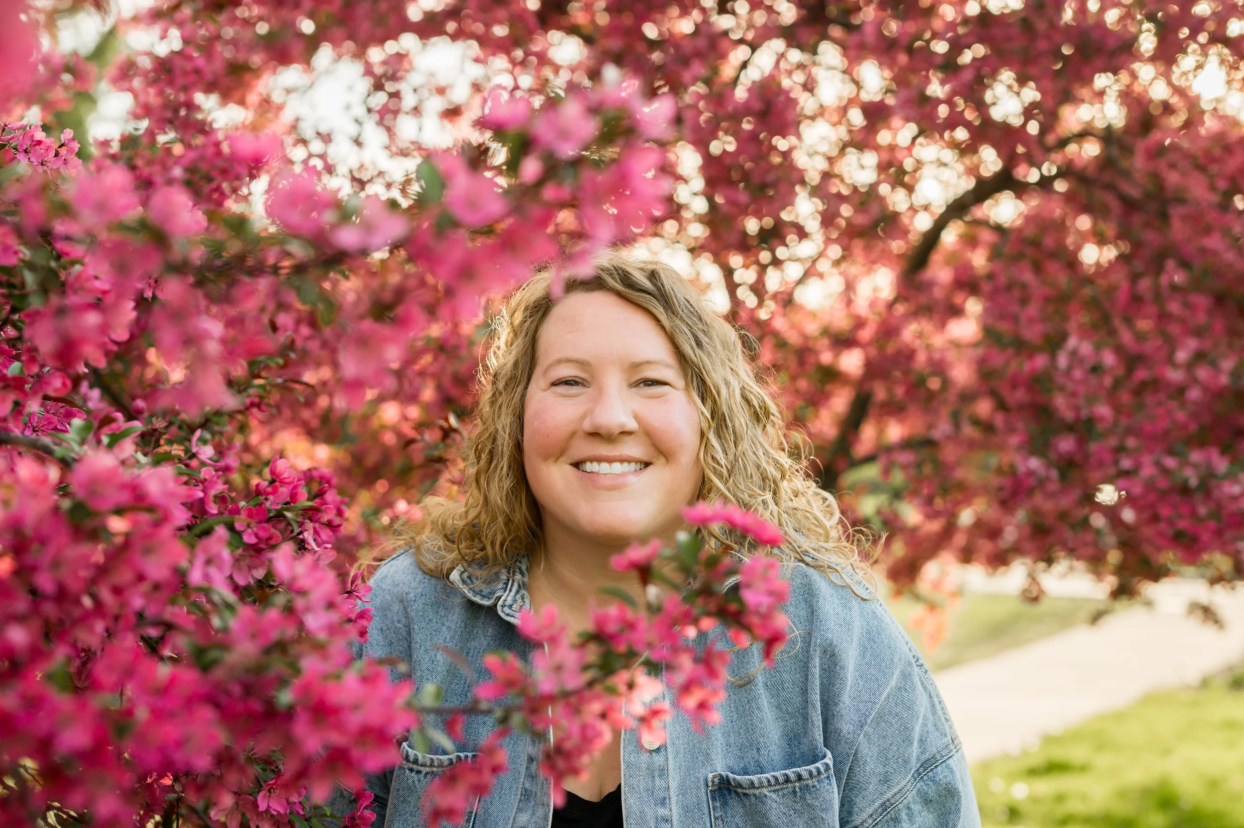 A woman with curly blonde hair smiling outdoors among pink flowering trees.