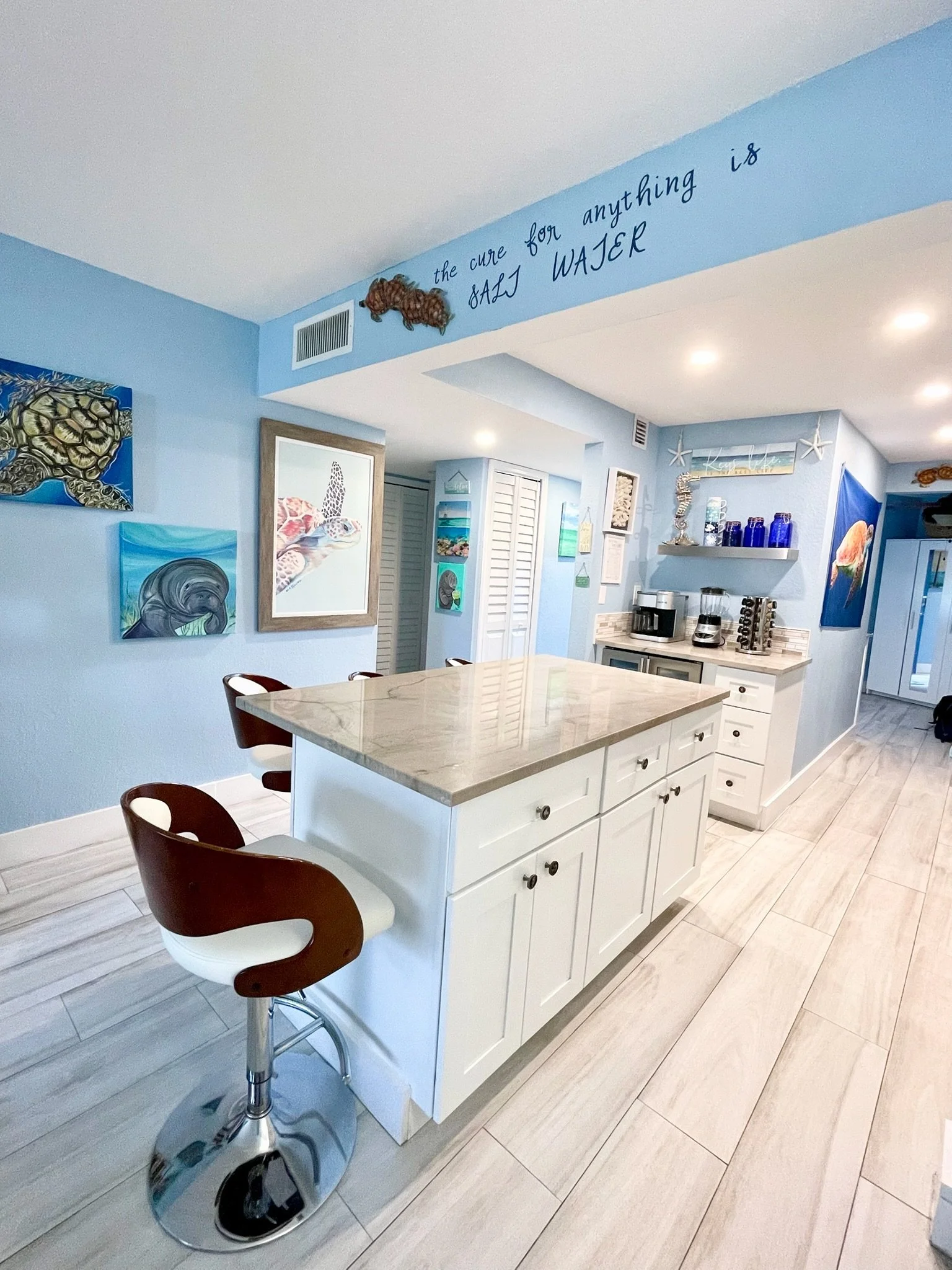 Bright kitchen with white island, brown and white bar stool, light blue walls decorated with ocean-themed art, and a coffee station with jars and starfish.