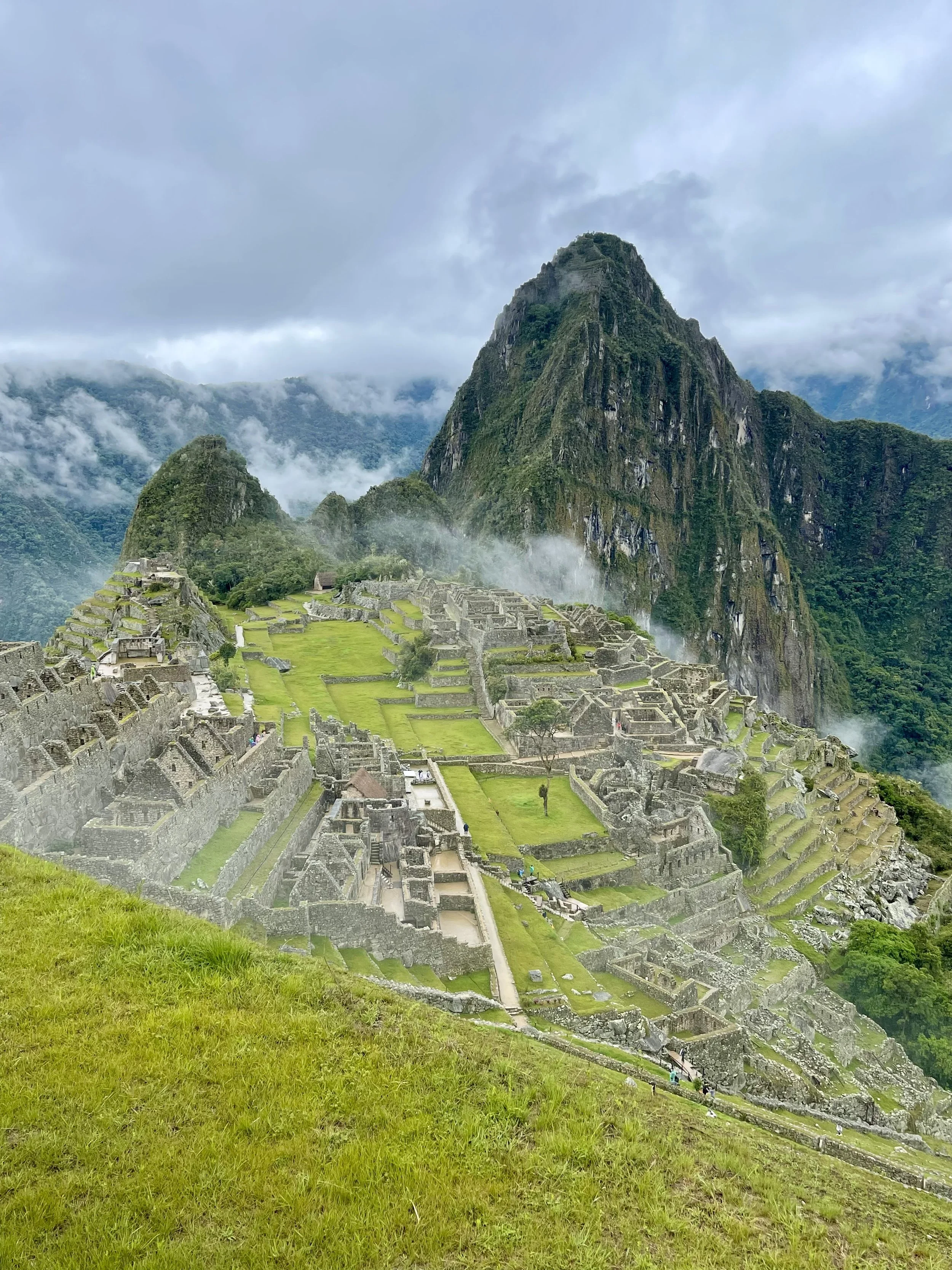 Ancient Inca ruins of Machu Picchu with lush green terraces and stone structures, surrounded by fog-covered mountains under a cloudy sky.