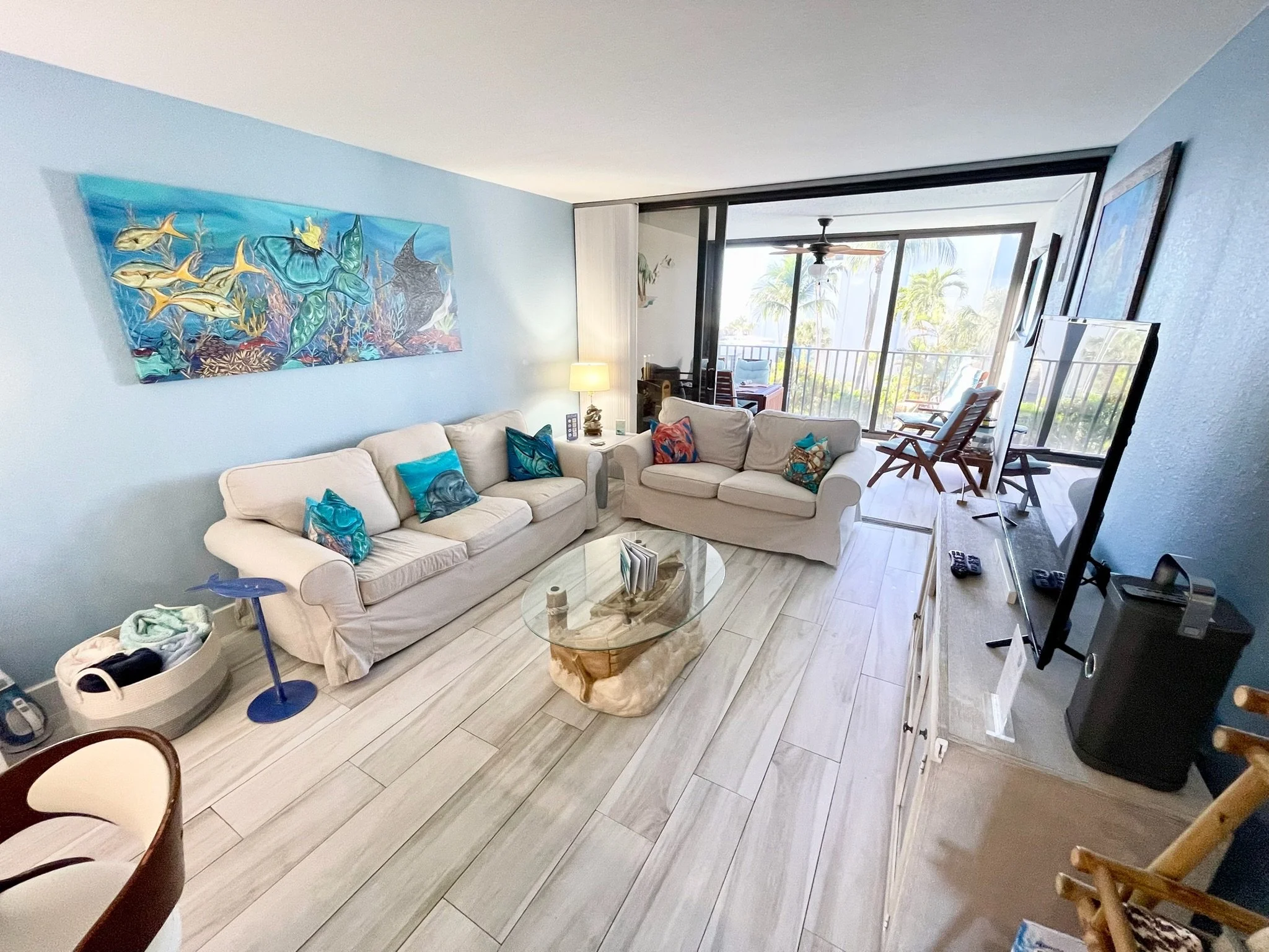 Living room with light blue walls, beige sofas with colorful pillows, a wooden coffee table, a lamp, a large painting of ocean fish on the wall, and a sliding glass door leading to a balcony with palm trees outside.