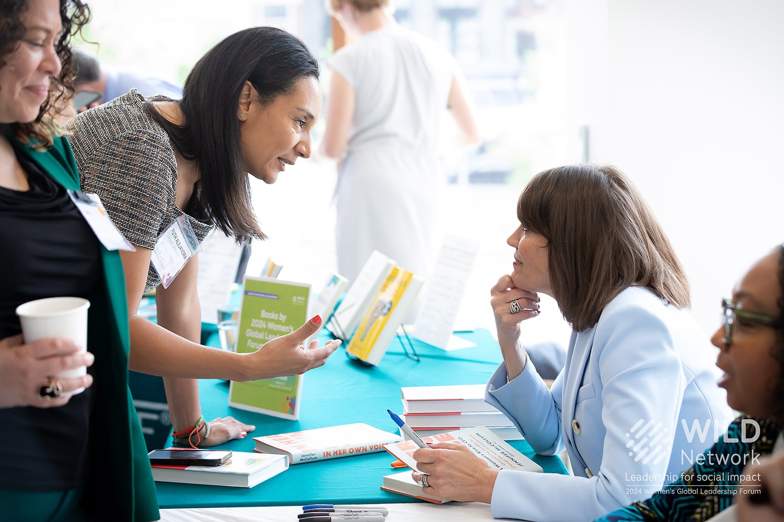 Women engaging in conversation at a book signing event, with books on the table and a bright, well-lit room.