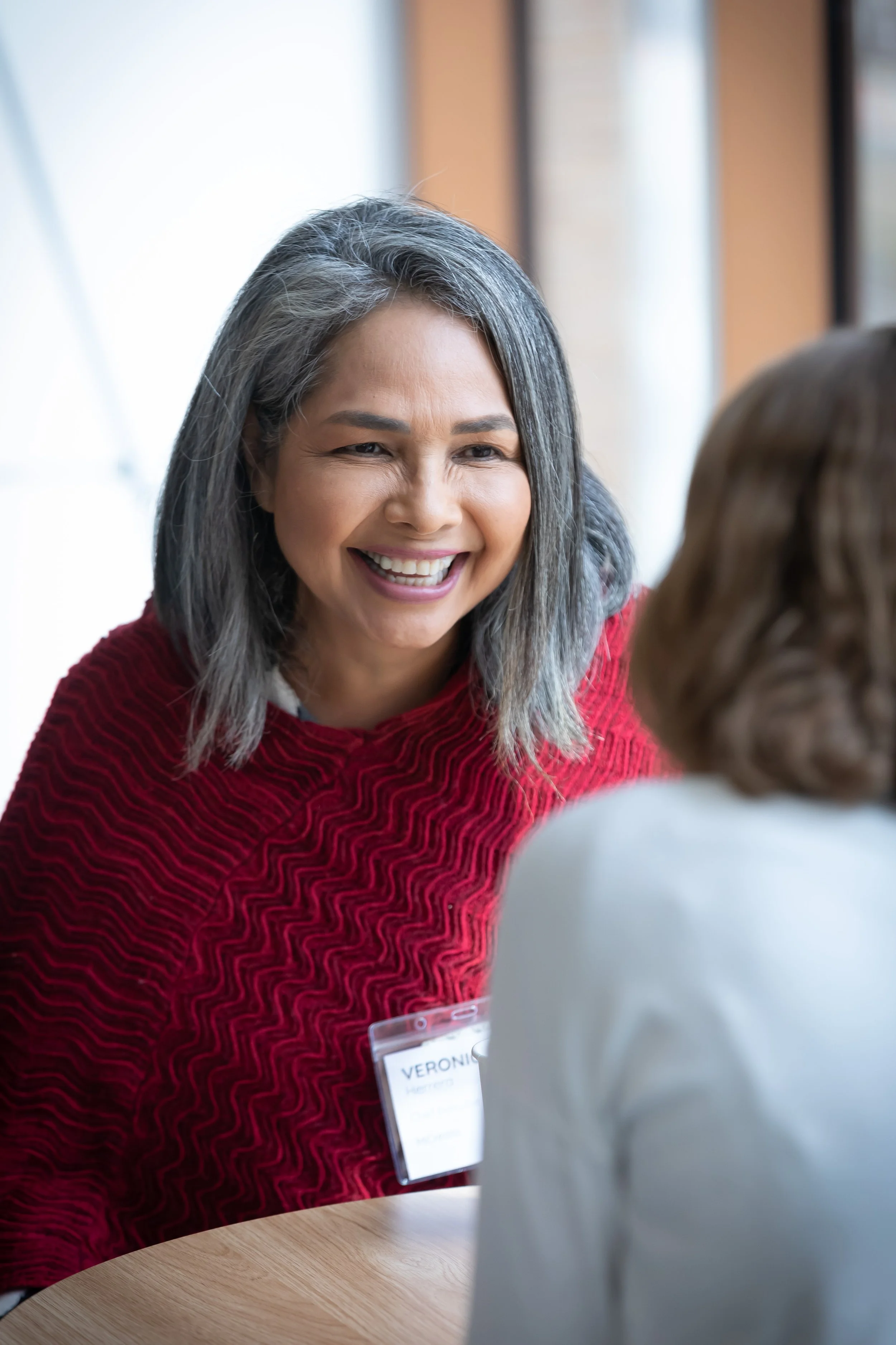 A middle-aged woman with gray hair, smiling warmly, engaged in conversation with another person at a table in a well-lit indoor setting.