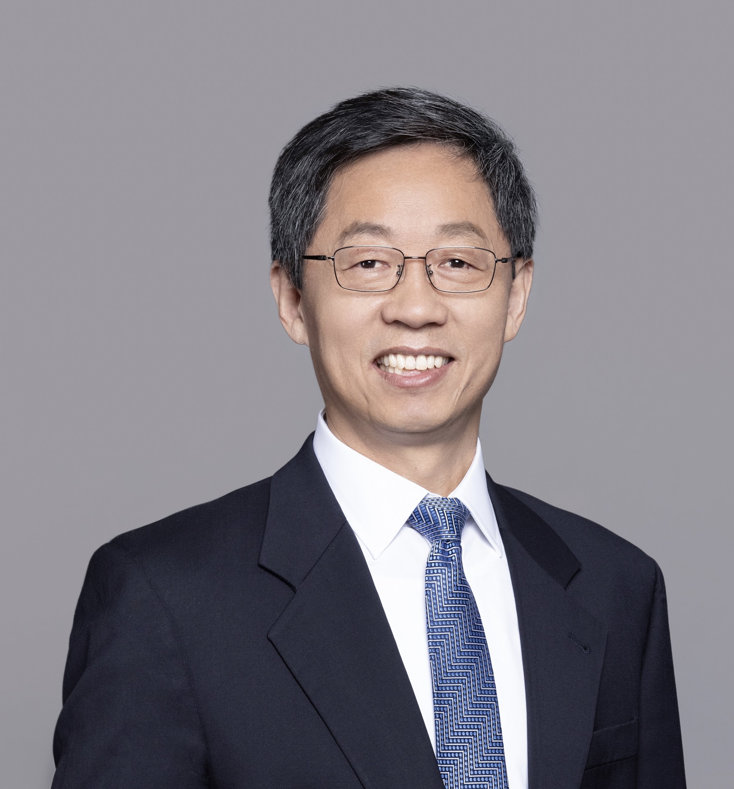 Portrait of a middle-aged Asian man wearing glasses, a black suit, white shirt, and a blue patterned tie, smiling against a plain gray background.