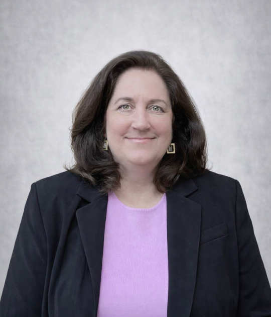 A woman with brown hair, wearing a black blazer and a lavender top, smiling at the camera, with a light gray background.