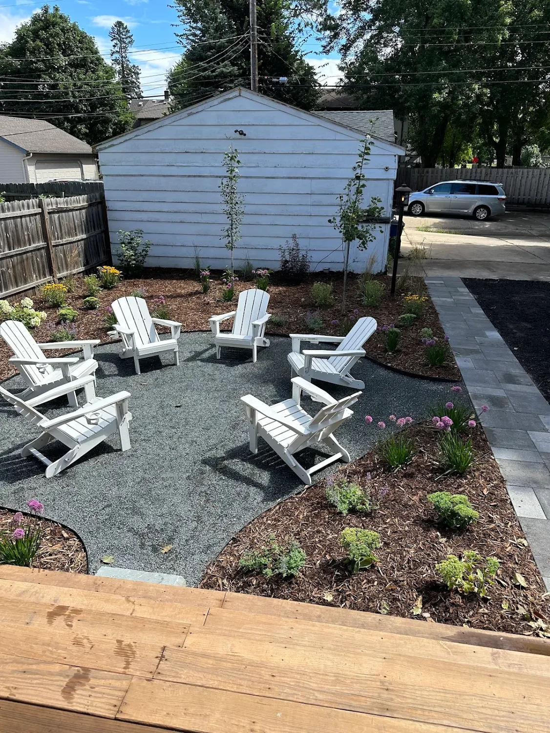 Backyard patio with six white Adirondack chairs arranged in a circle on a gravel surface. Surrounding the gravel area are flower beds with yellow and purple flowers, small shrubs, and young trees. There is a wooden fence on the left side, a small white shed in the background, and a paved walkway on the right side leading to a street with a parked silver minivan. Overhead power lines are visible, and the sky is partly cloudy.