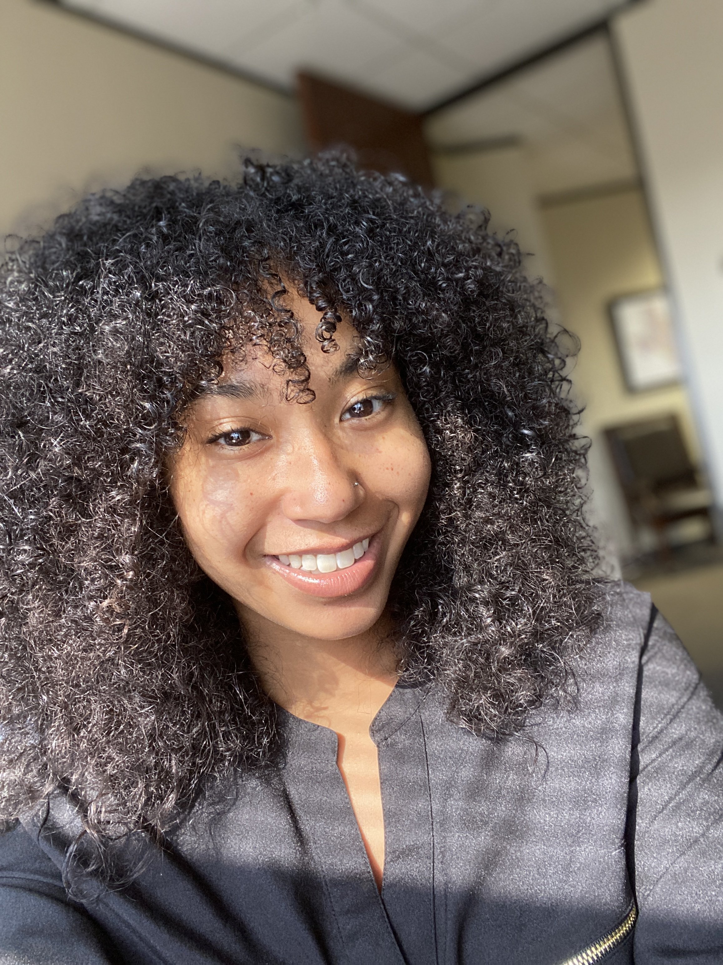 A young woman with curly black hair taking a selfie indoors, smiling.