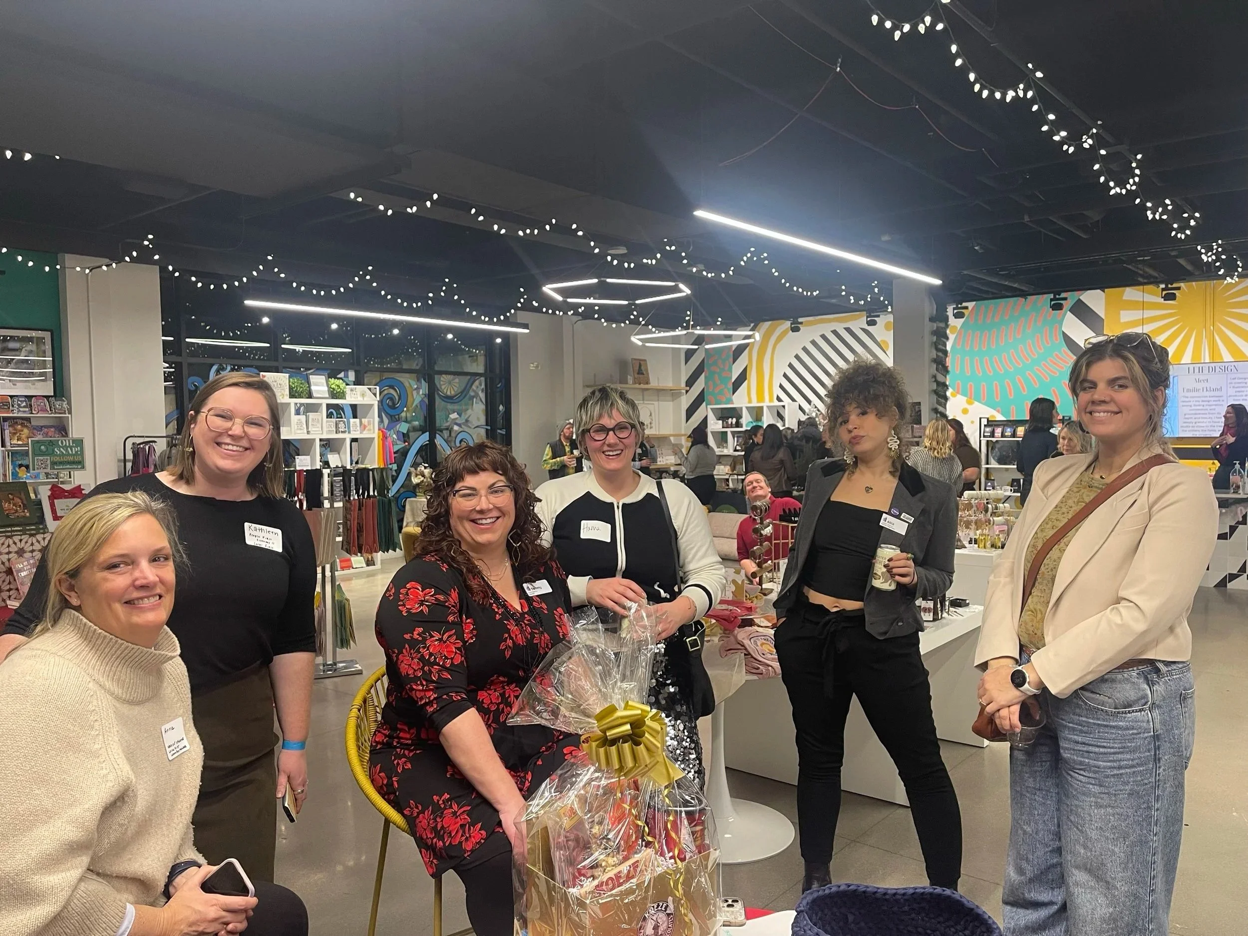 Group of women at an event, some smiling and holding gifts, in a decorated indoor space with string lights and colorful wall art.