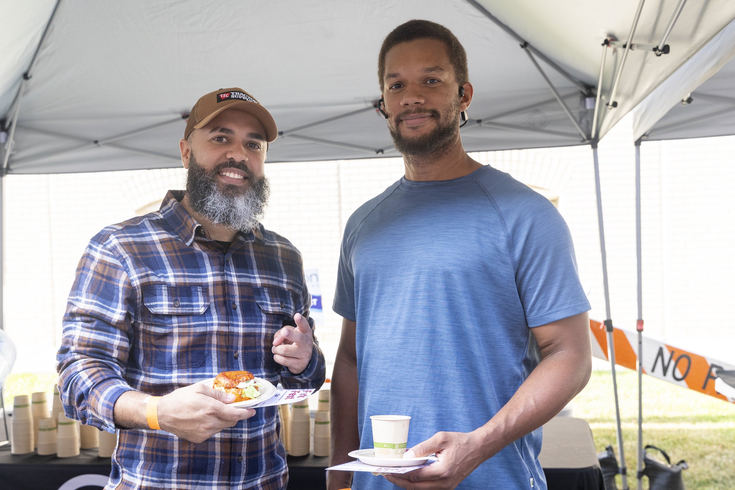 Two men at an outdoor event under a tent. One man with a beard, plaid shirt, and cap holds a plate with food and smiles. The other man with short hair and a blue shirt holds a cup and plate, with a slight smile. There are stacked cups and a sign in the background.