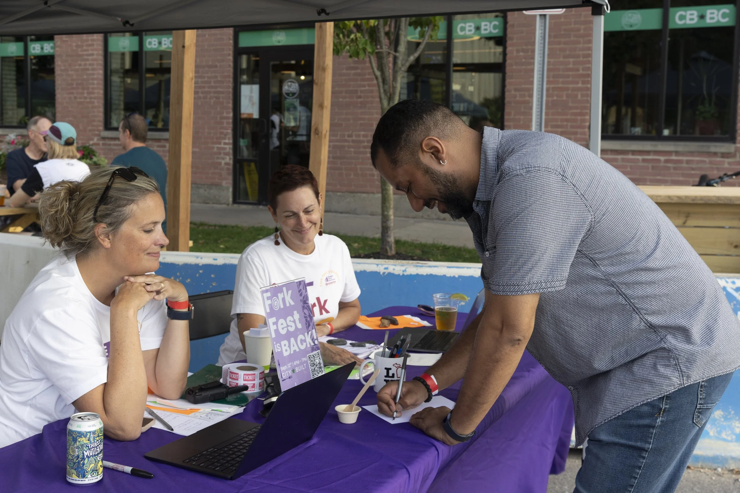 People at an outdoor event registration table, with two women seated behind the table and a man standing, filling out a form. The table has a purple tablecloth, laptops, pens, and a sign that reads 'Fork Fest is Back!' in front of the women. There are other people sitting at tables and a brick building with large windows in the background.