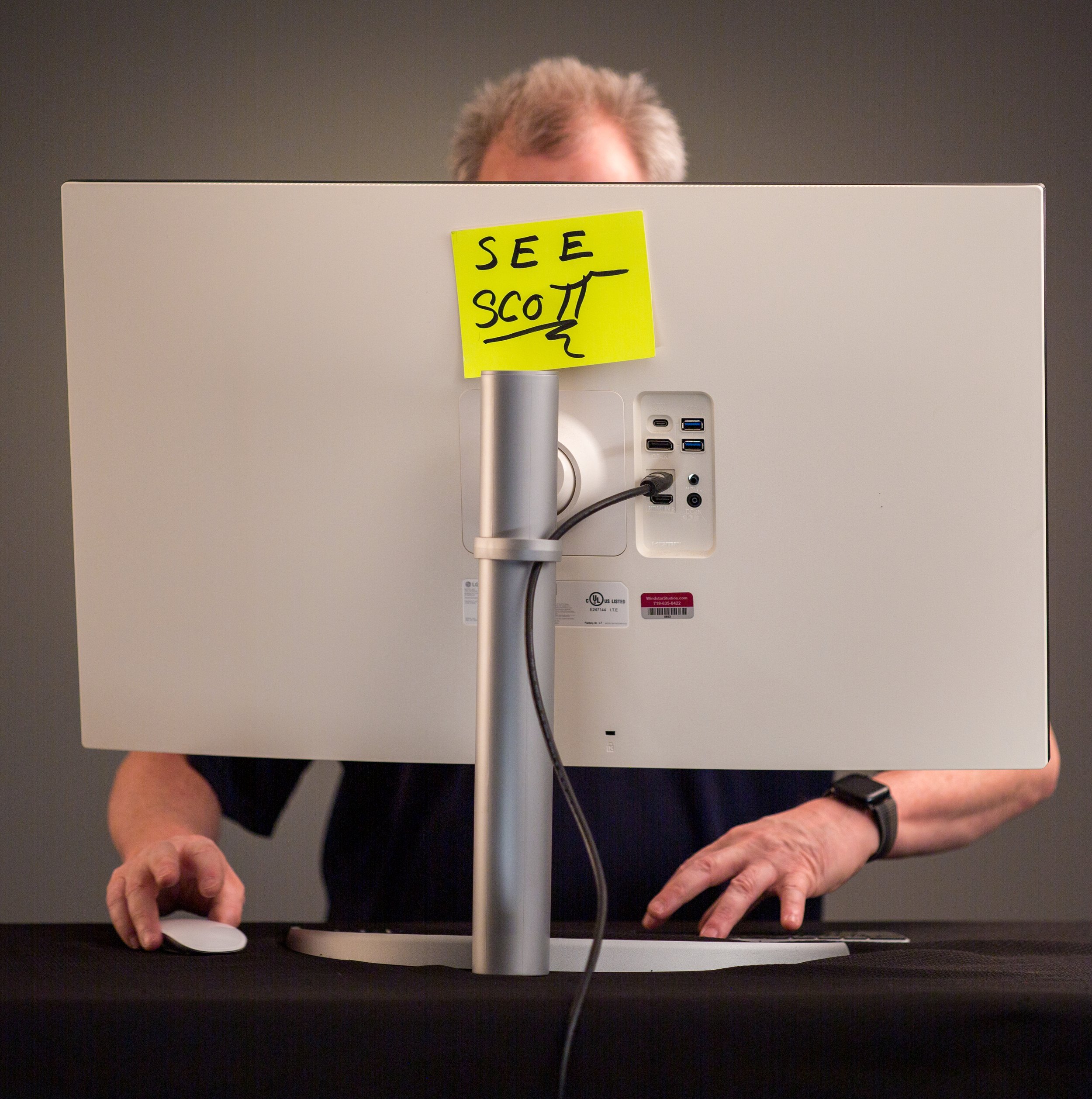 Person working at a computer with a yellow sticky note on the monitor that says 'SEE SCOTT'.