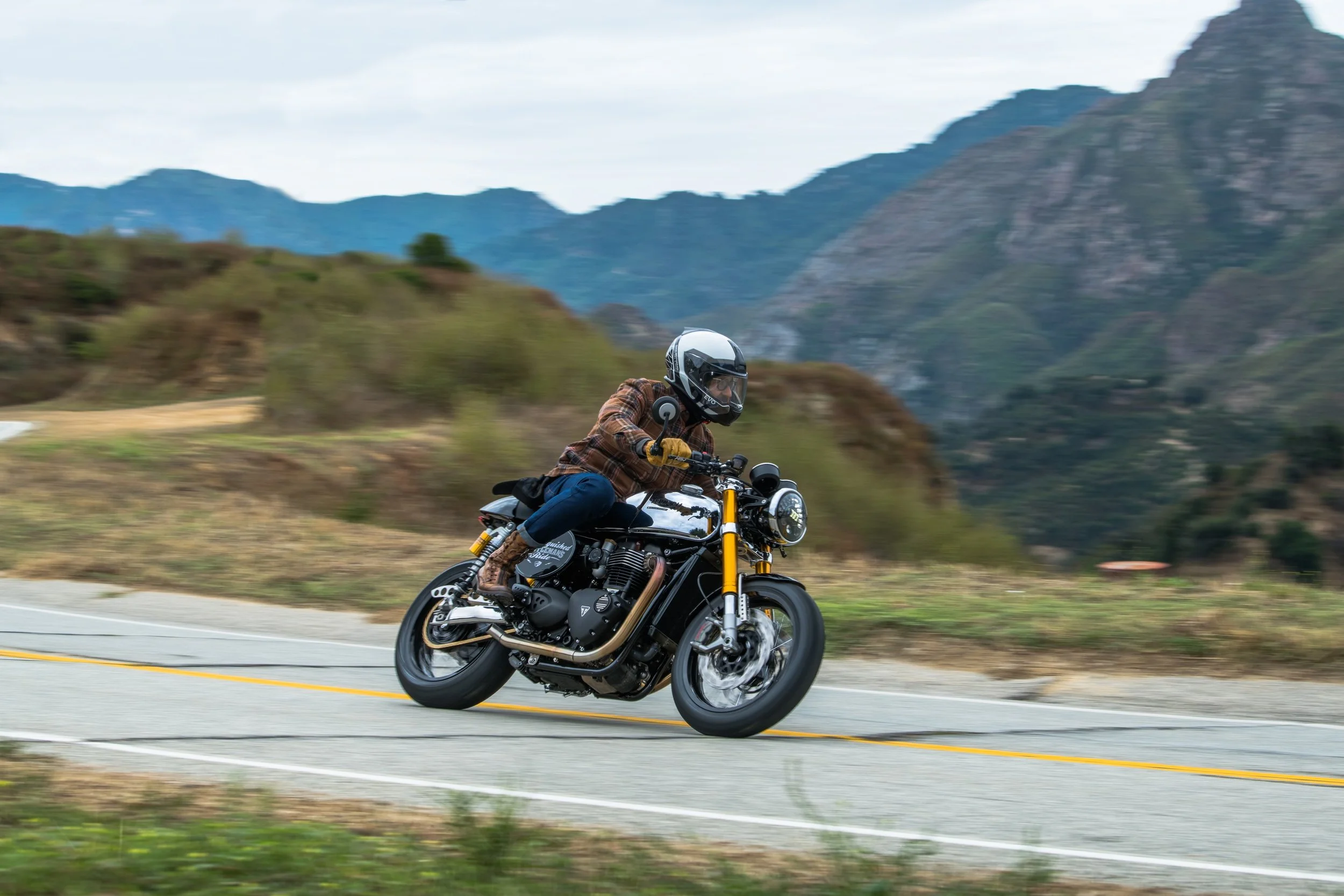 A person riding a black motorcycle on a winding mountain road, wearing a helmet, plaid jacket, and riding boots, with a mountainous landscape in the background.