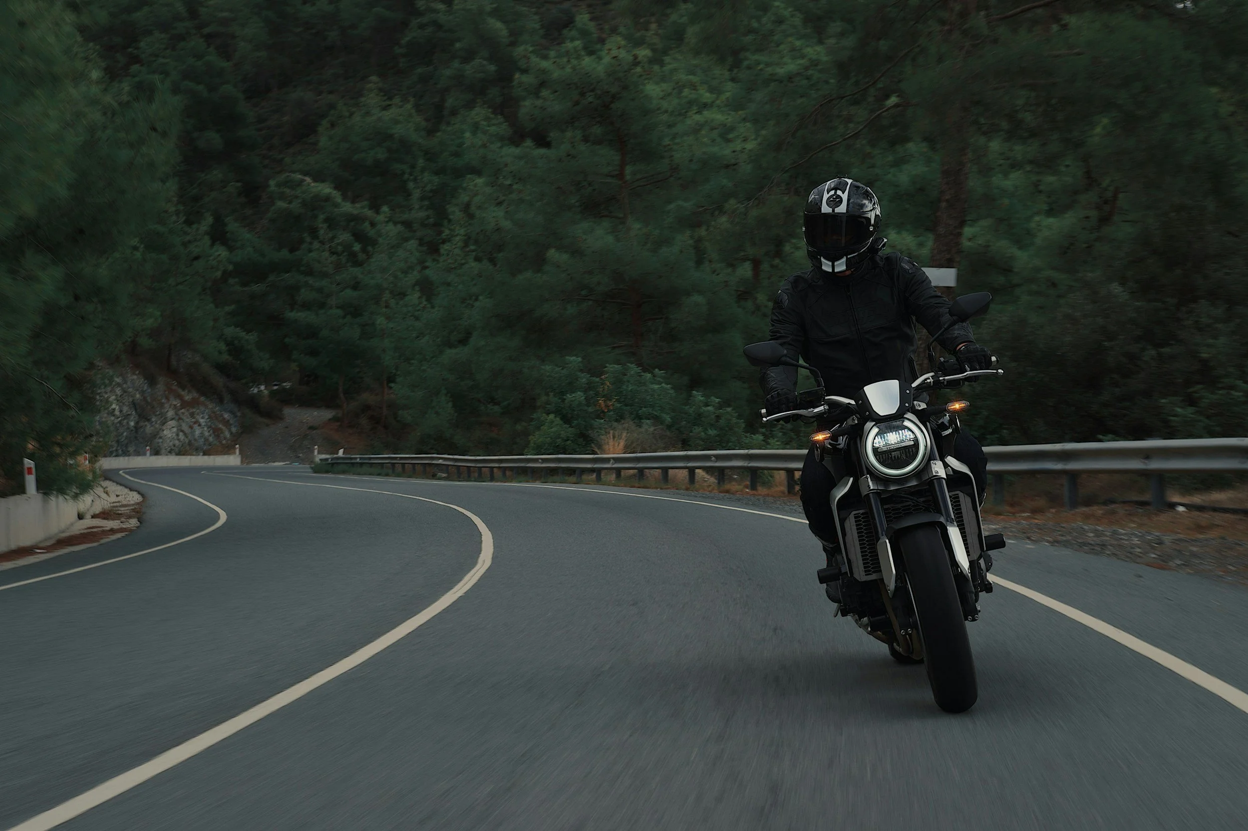 Motorcyclist wearing black protective gear and helmet riding on a winding mountain road surrounded by green trees.