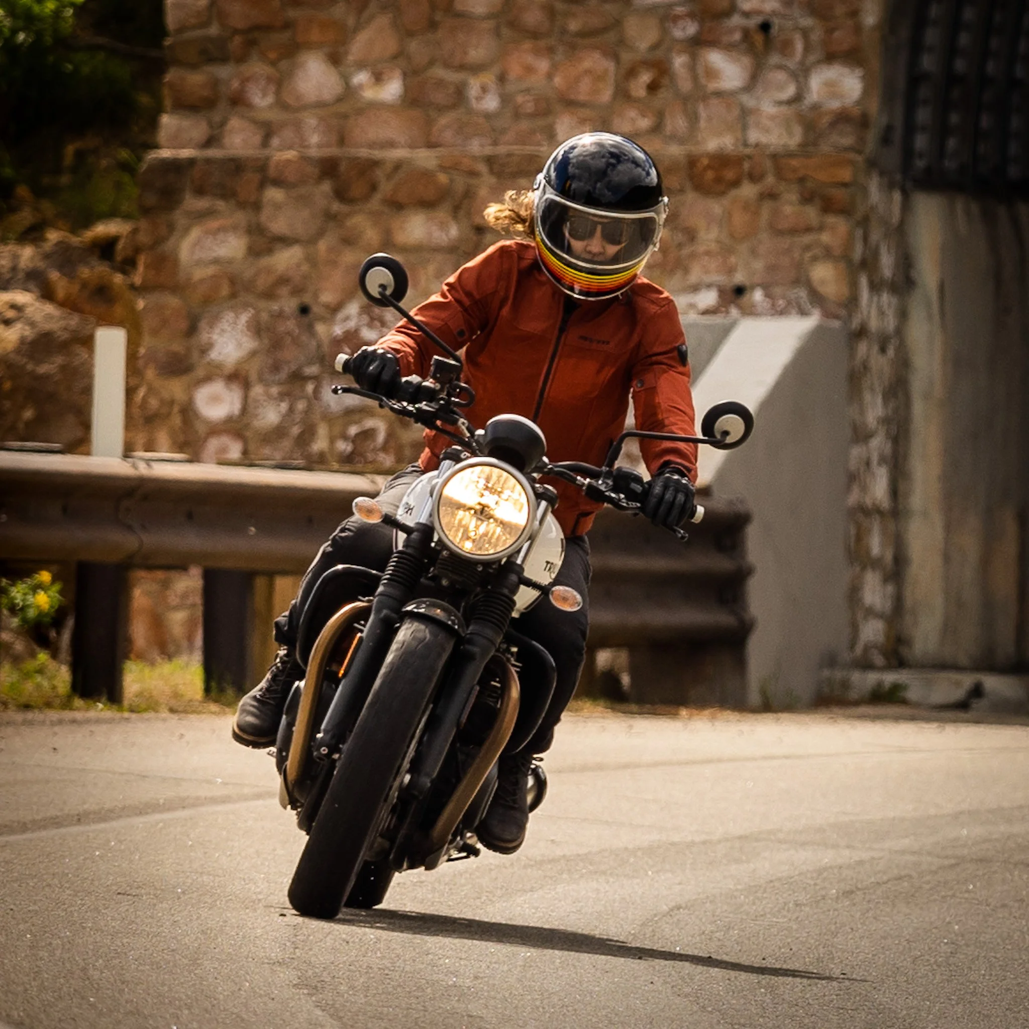 A person riding a motorcycle on a curved road, wearing a helmet with a visor, a brown jacket, and black gloves, with a stone wall and guardrail in the background.