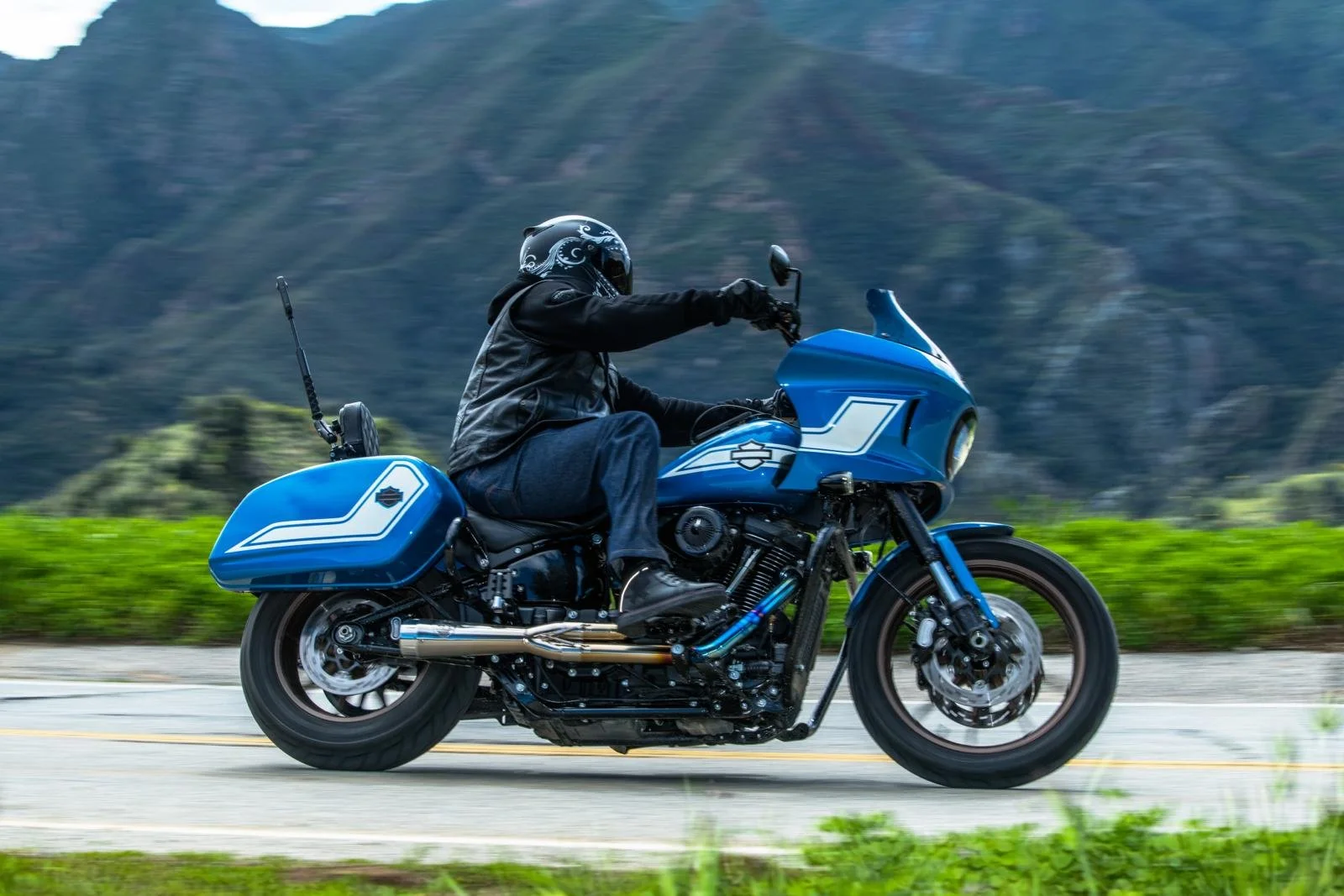 A person riding a blue motorcycle on a scenic road with mountains in the background.