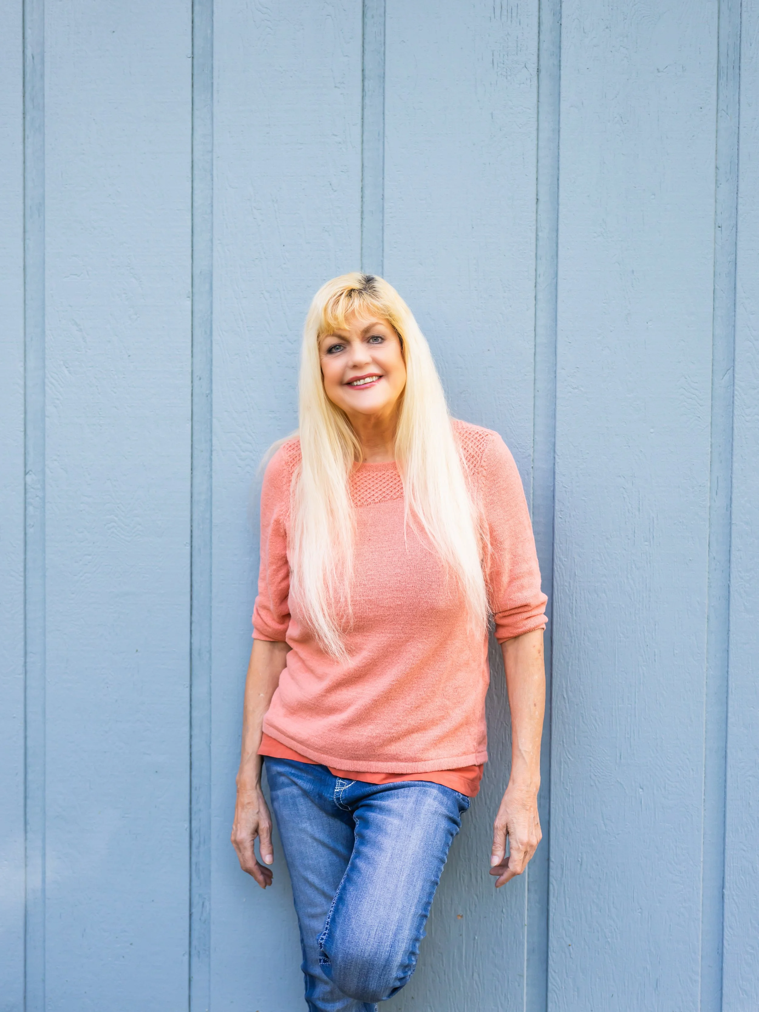 A woman with long blonde hair, wearing a pink sweater and blue jeans, leaning against a light blue wooden wall, smiling.