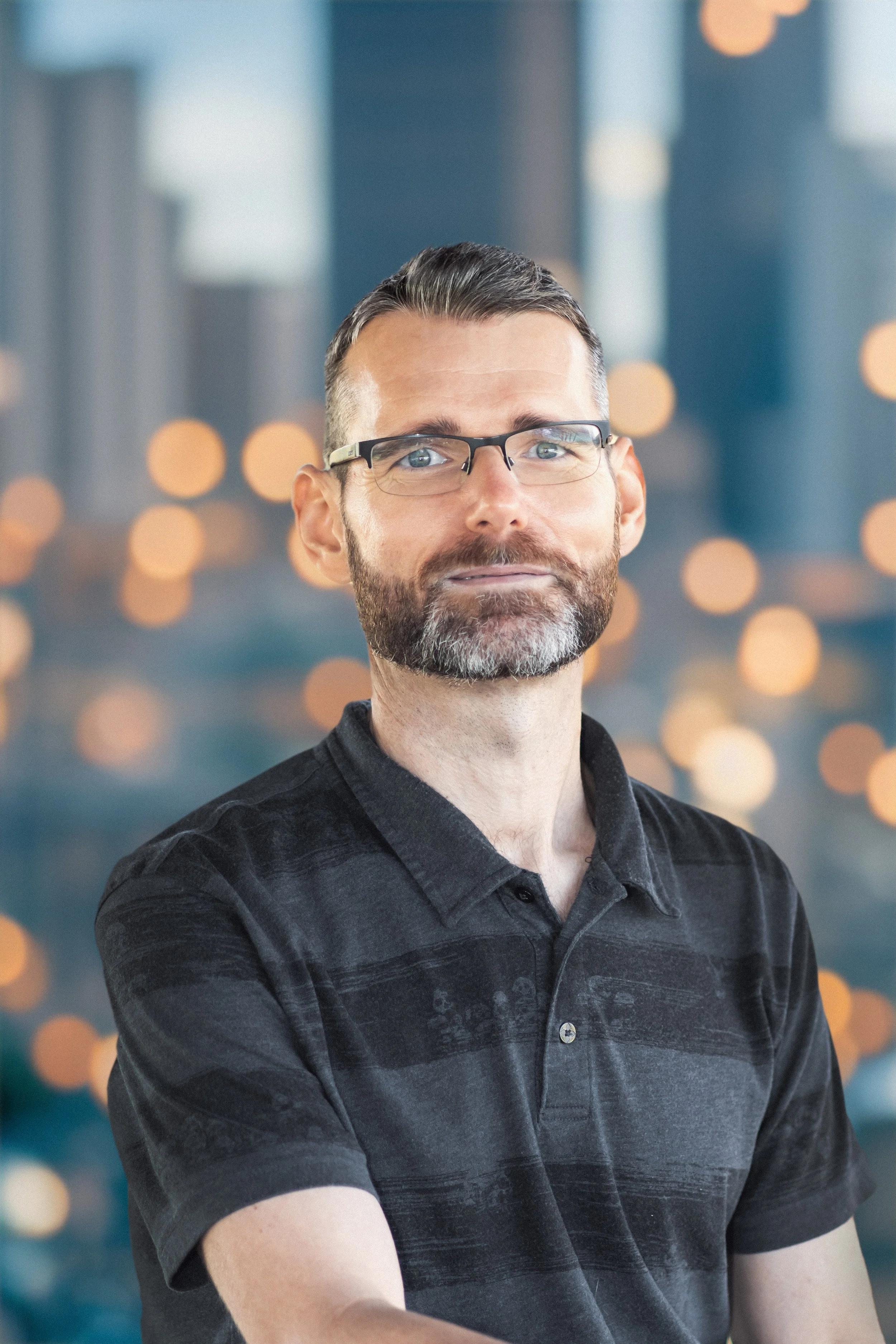 A man with glasses, short dark hair, and a beard, wearing a black polo shirt, standing in front of a blurred cityscape with bokeh lights.