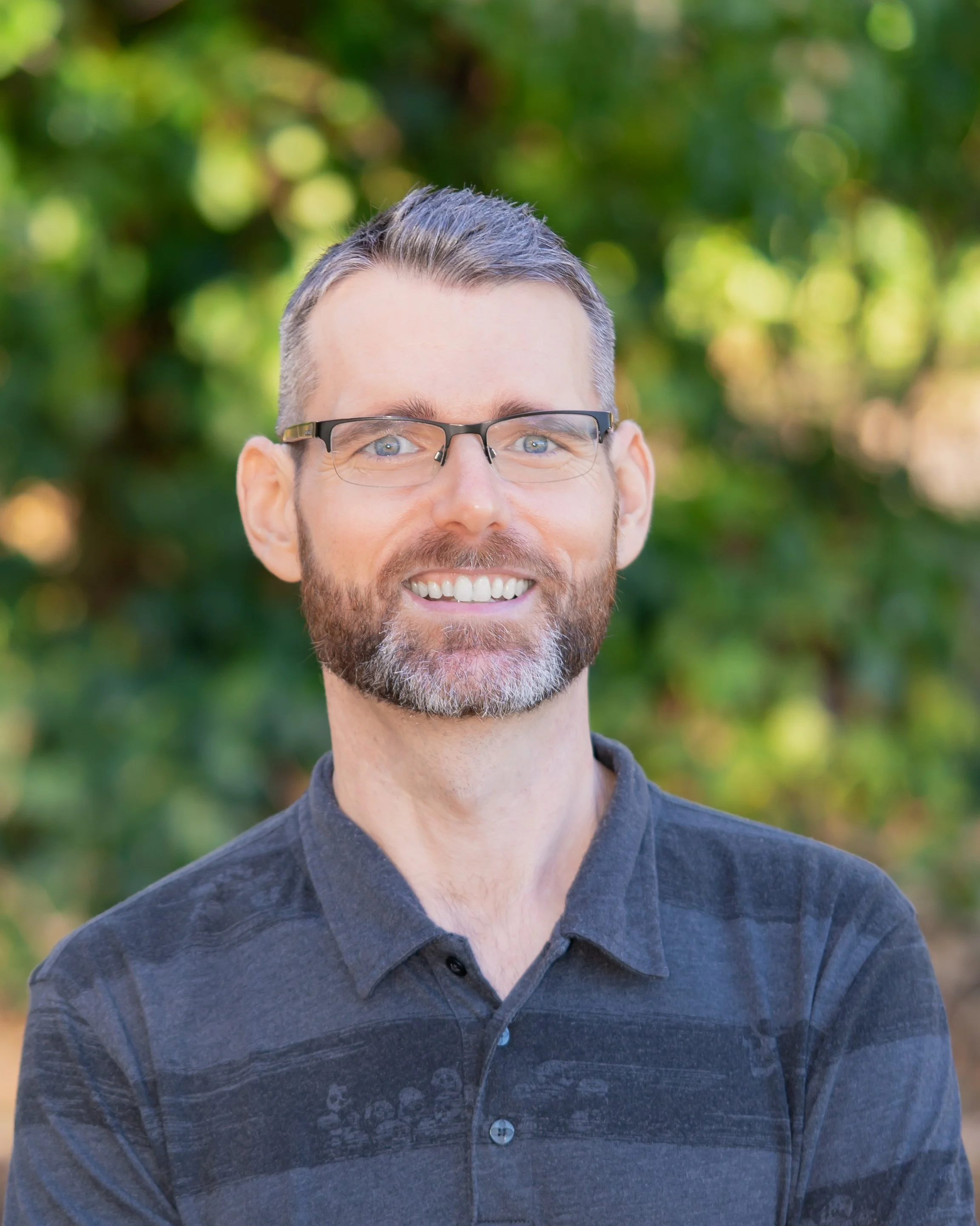 A smiling middle-aged man with glasses and a beard, wearing a dark polo shirt, standing outdoors with green trees in the background.