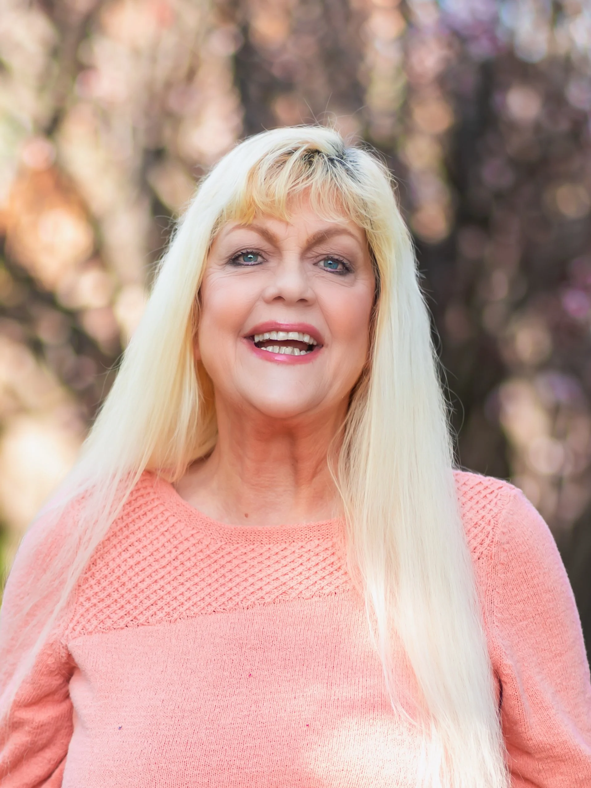 A smiling blonde woman with blue eyes wearing a peach-colored sweater outdoors with pink and brown blurred trees in the background.