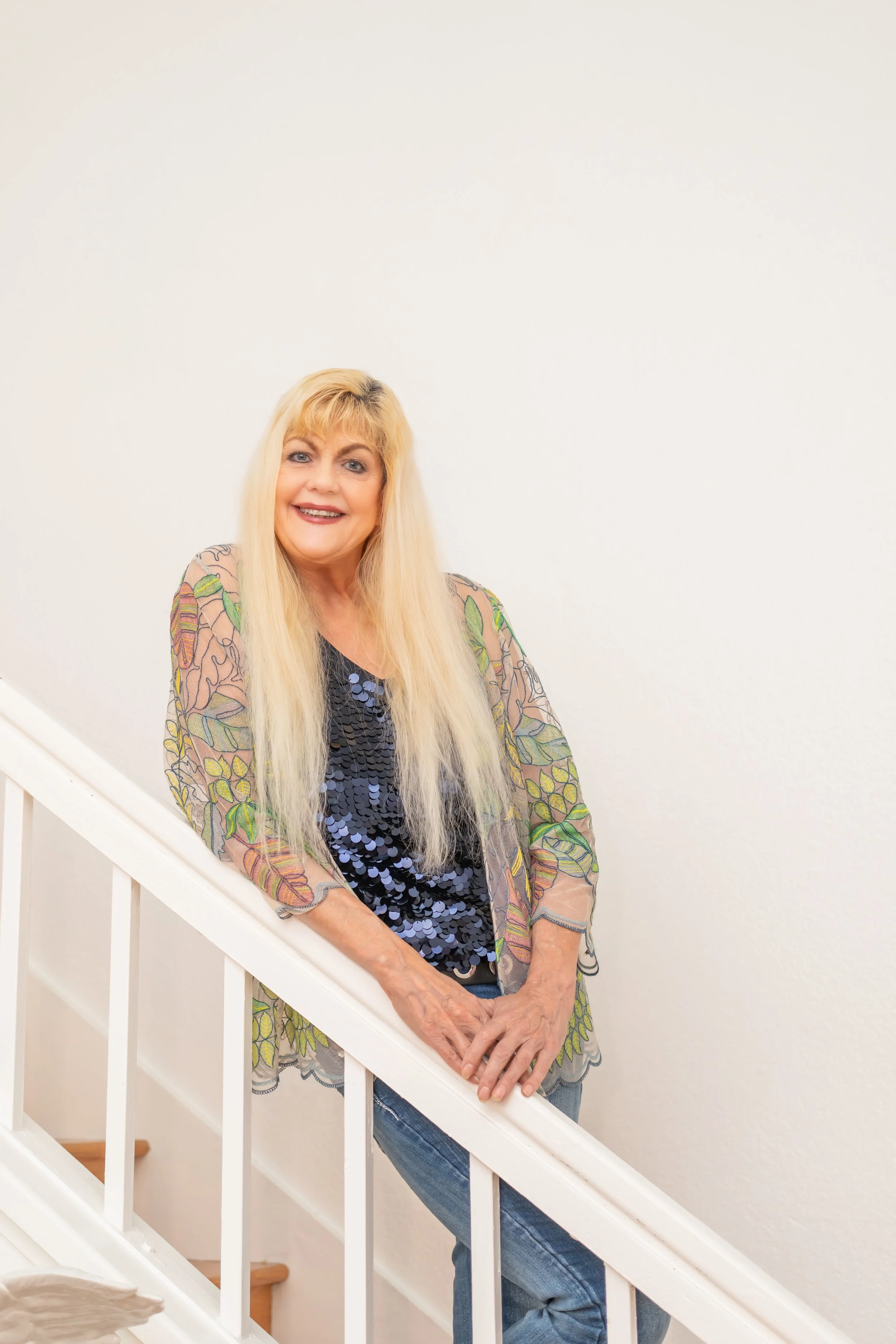 A smiling woman with long blonde hair standing on a staircase, wearing a colorful, sheer, floral-patterned jacket over a sequined top and blue jeans.