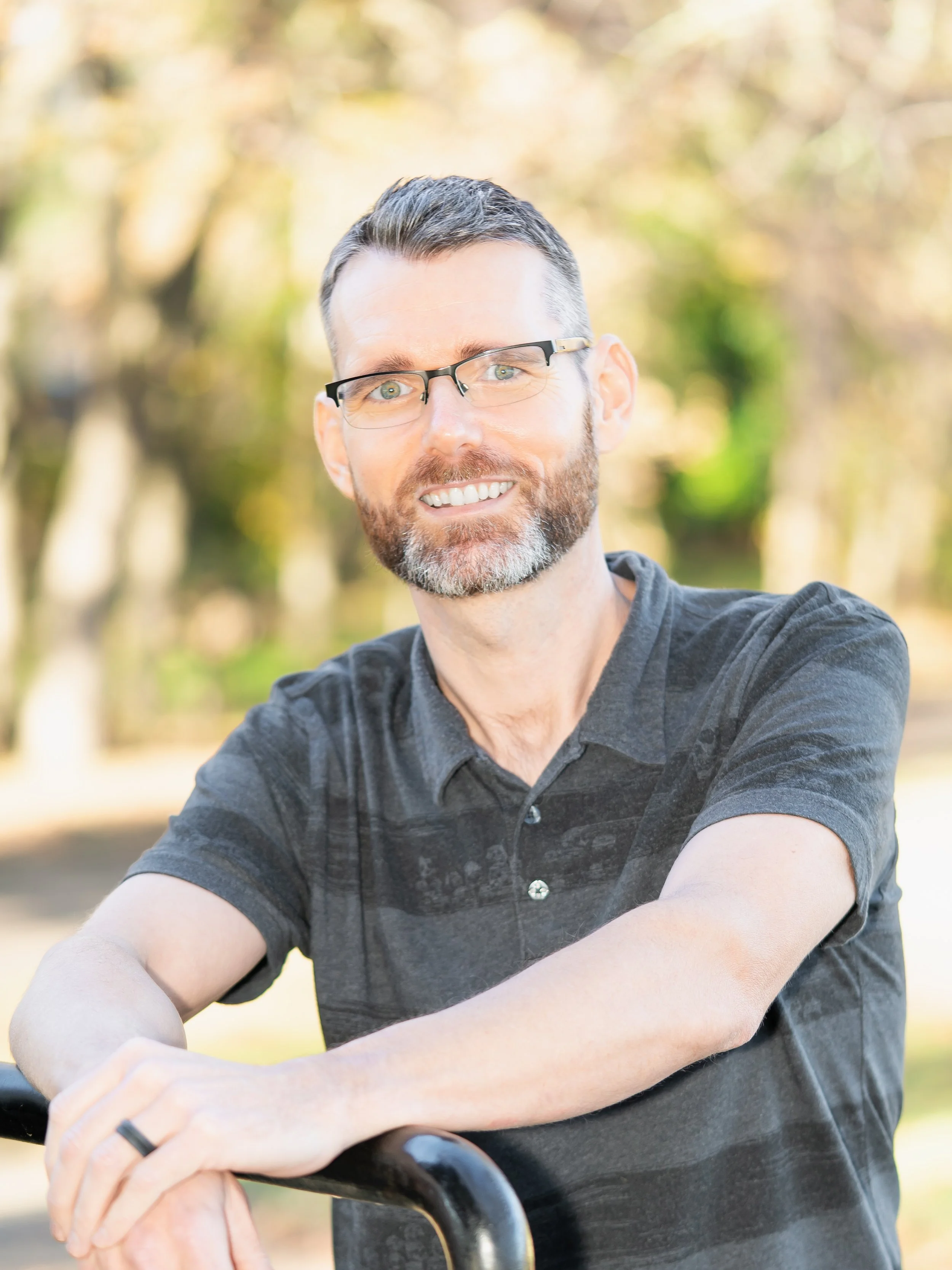 A man with glasses and a beard, smiling and leaning on a bicycle handlebar outdoors, with trees in the background.