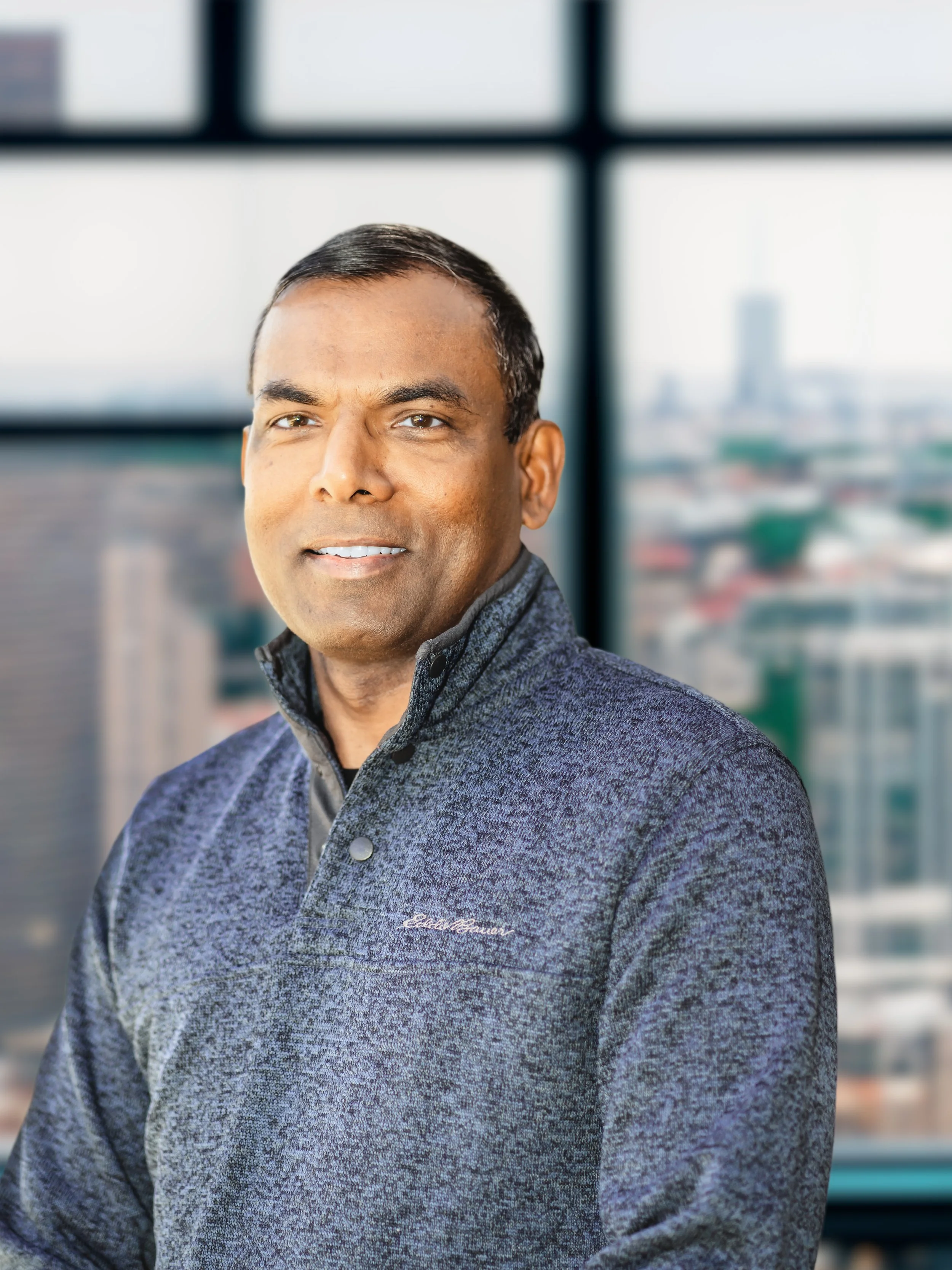 Professional man with short black hair in a grey fleece sweater, standing in front of a large window overlooking a cityscape.