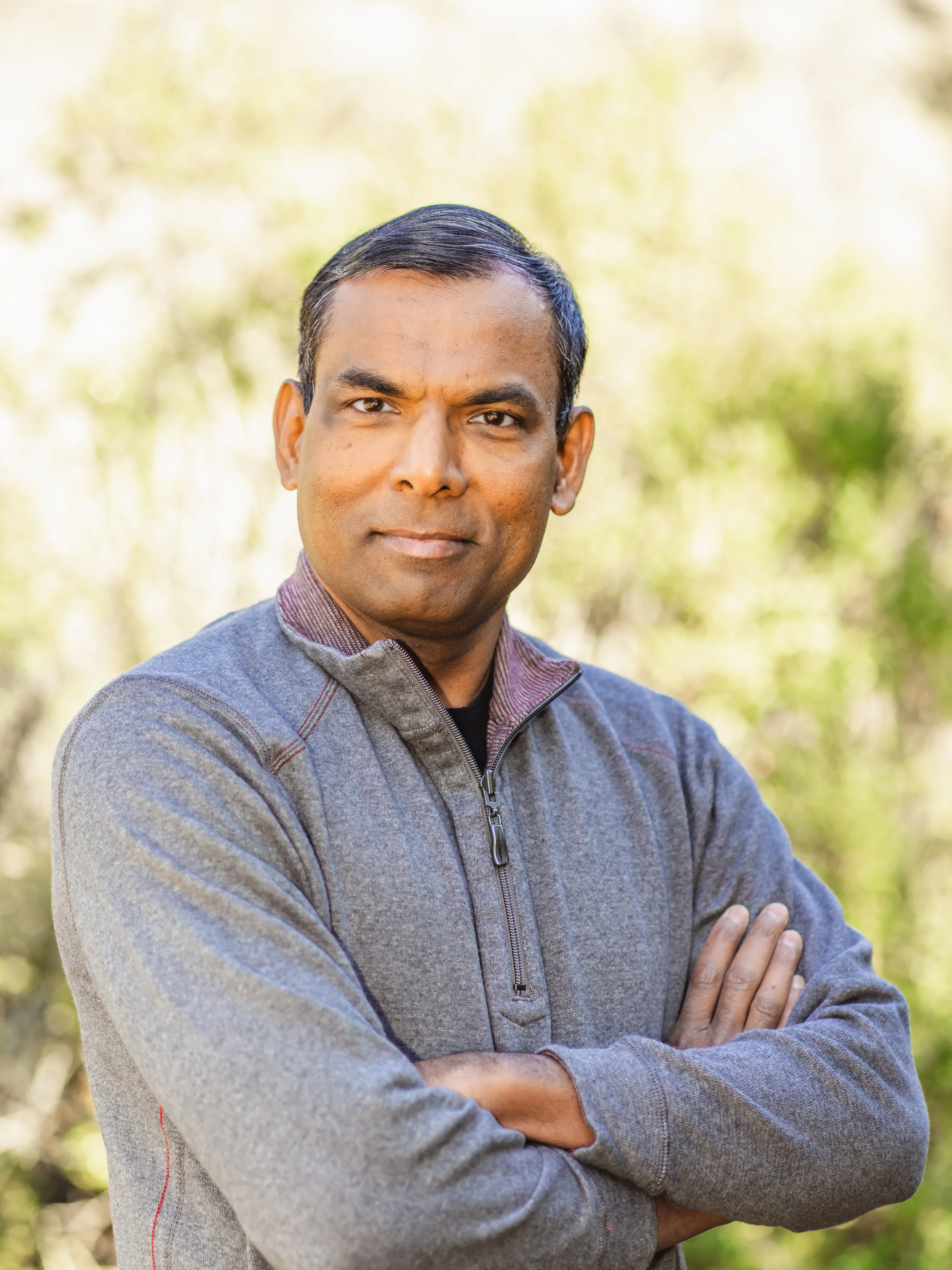 A man with short dark hair, wearing a gray zip-up jacket, standing outdoors with arms crossed and a neutral expression, with blurred greenery in the background.
