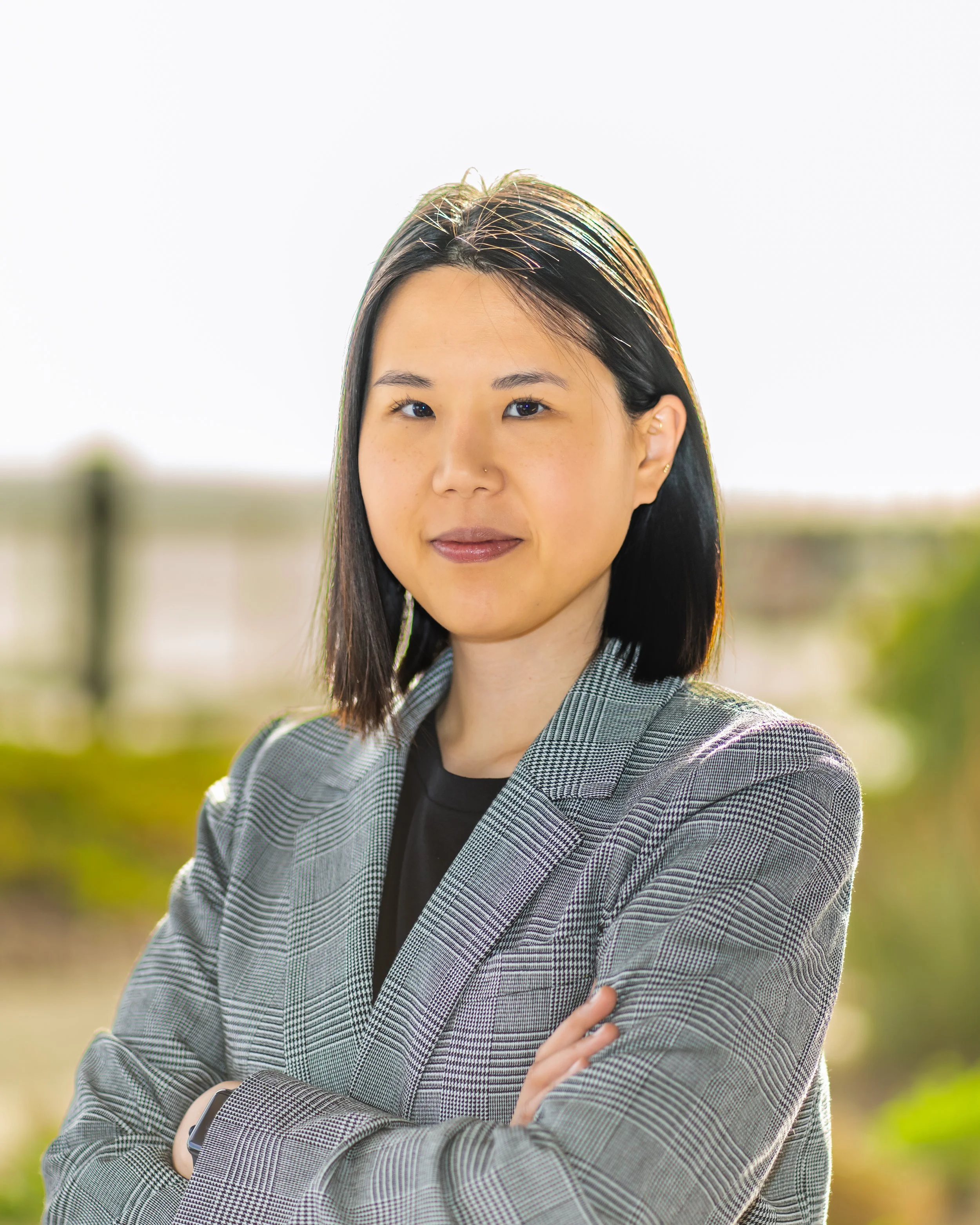 A woman with shoulder-length dark hair, wearing a checkered blazer and black shirt, standing outdoors with arms crossed and a blurred natural background.