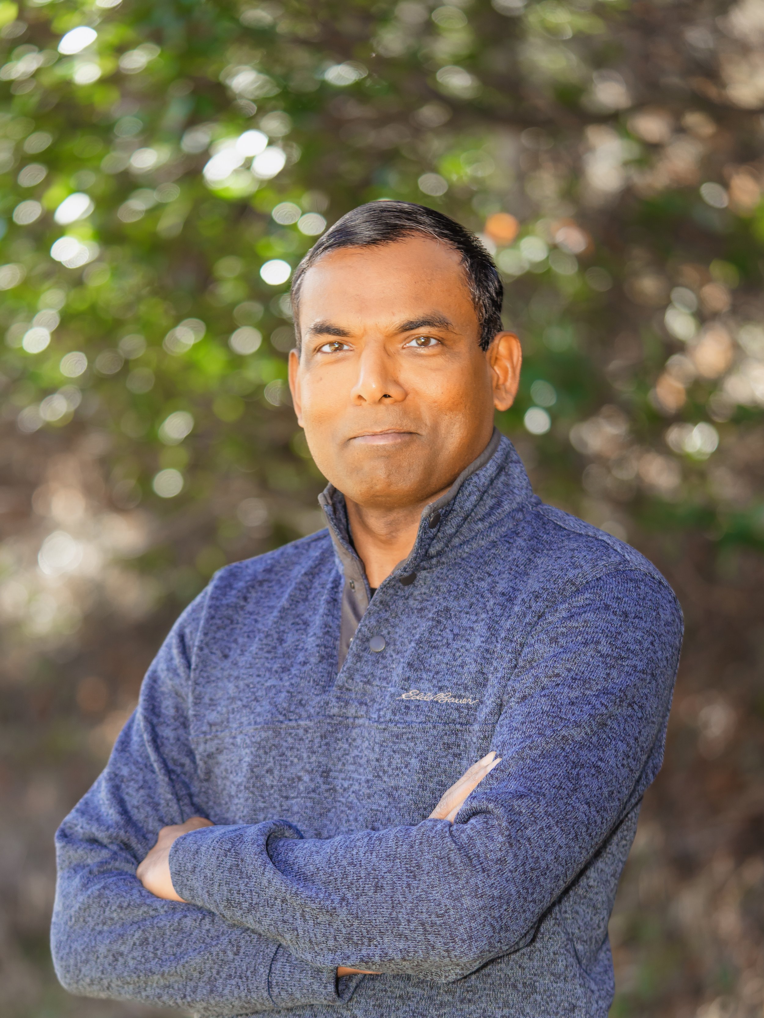 A man with short black hair and brown skin, wearing a blue sweater. He is standing outdoors with his arms crossed, looking directly at the camera. Blurred green foliage in the background.