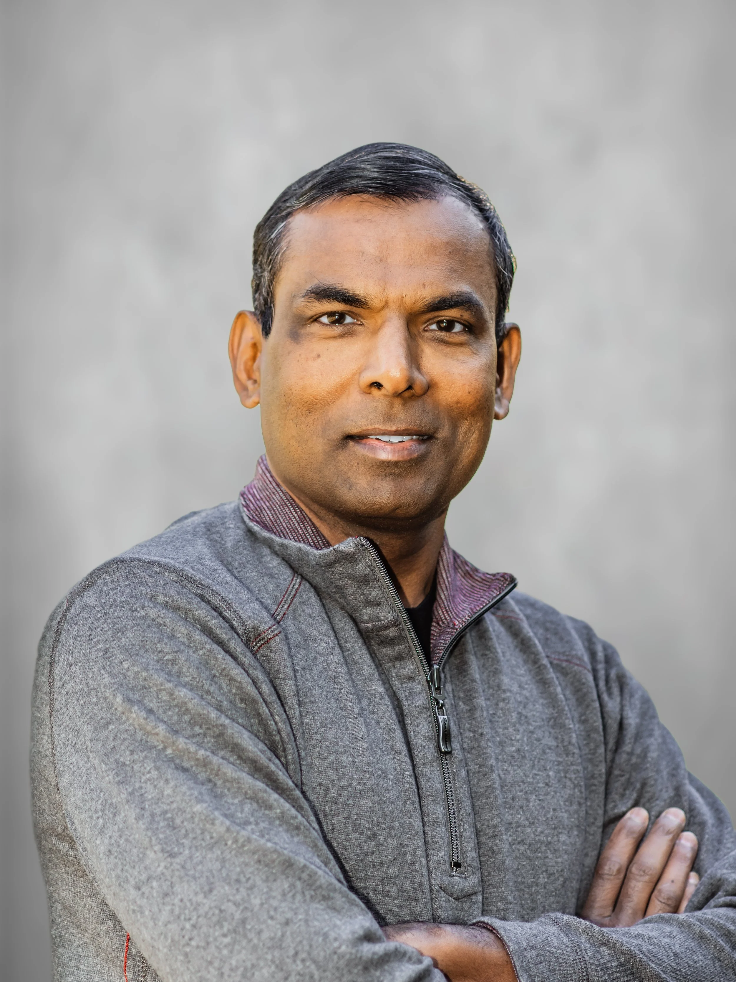 A man with dark hair and medium skin tone, wearing a gray zip-up jacket, crossing his arms, standing in front of a plain light gray background.