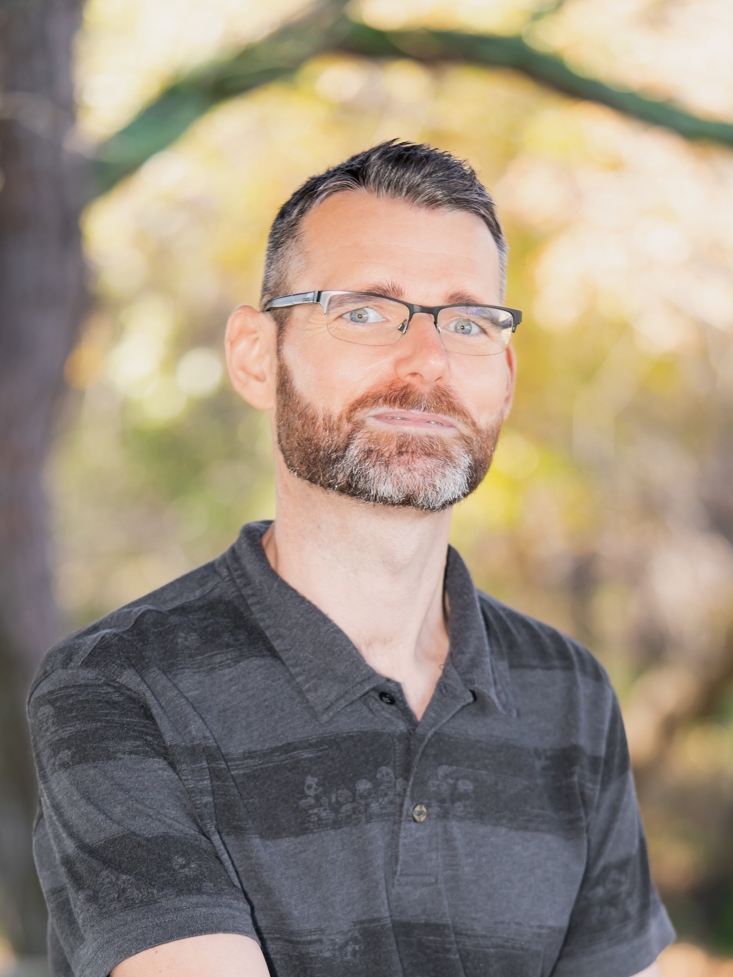 A man with short dark hair, a beard with gray, wearing glasses and a dark, patterned polo shirt, standing outdoors with autumn-colored trees in the background.
