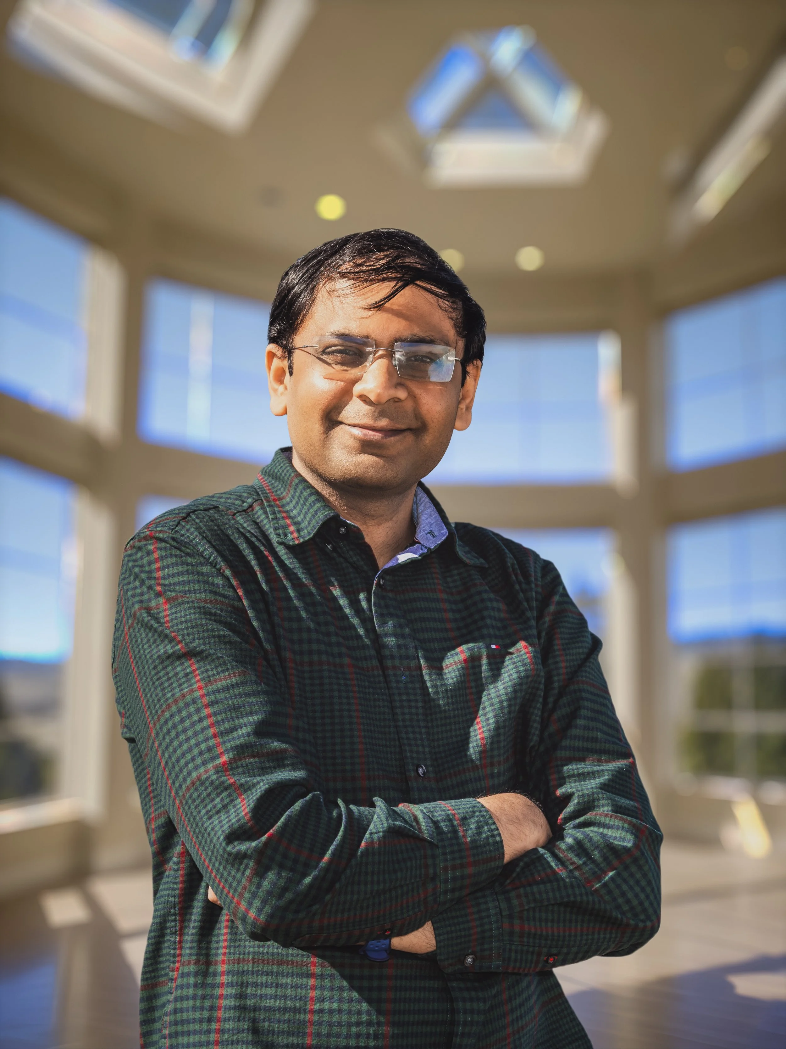 A man with glasses and a checkered shirt standing with arms crossed inside a bright room with large windows.