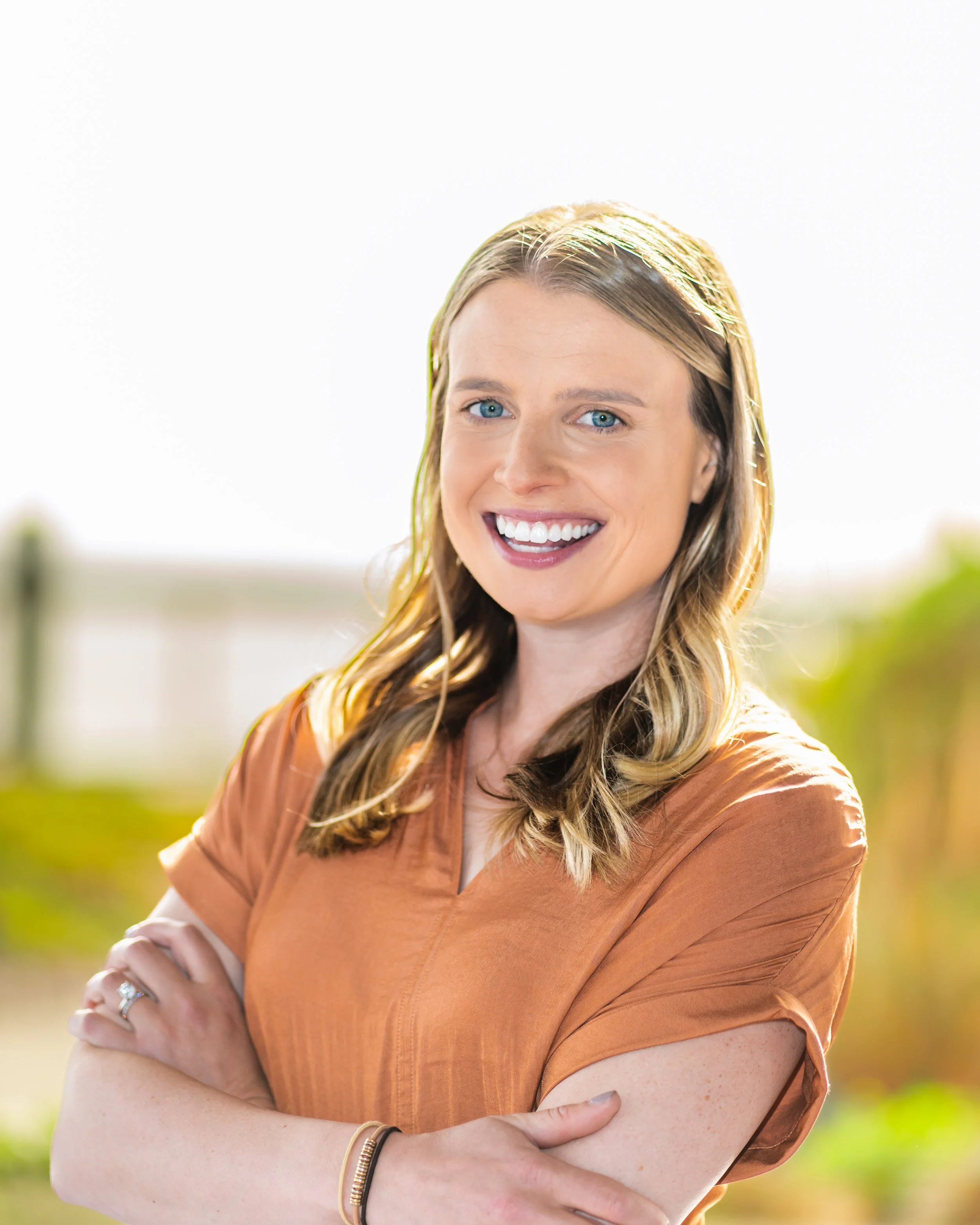 A smiling woman with blue eyes and long blonde hair standing outdoors with arms crossed, wearing an orange top.
