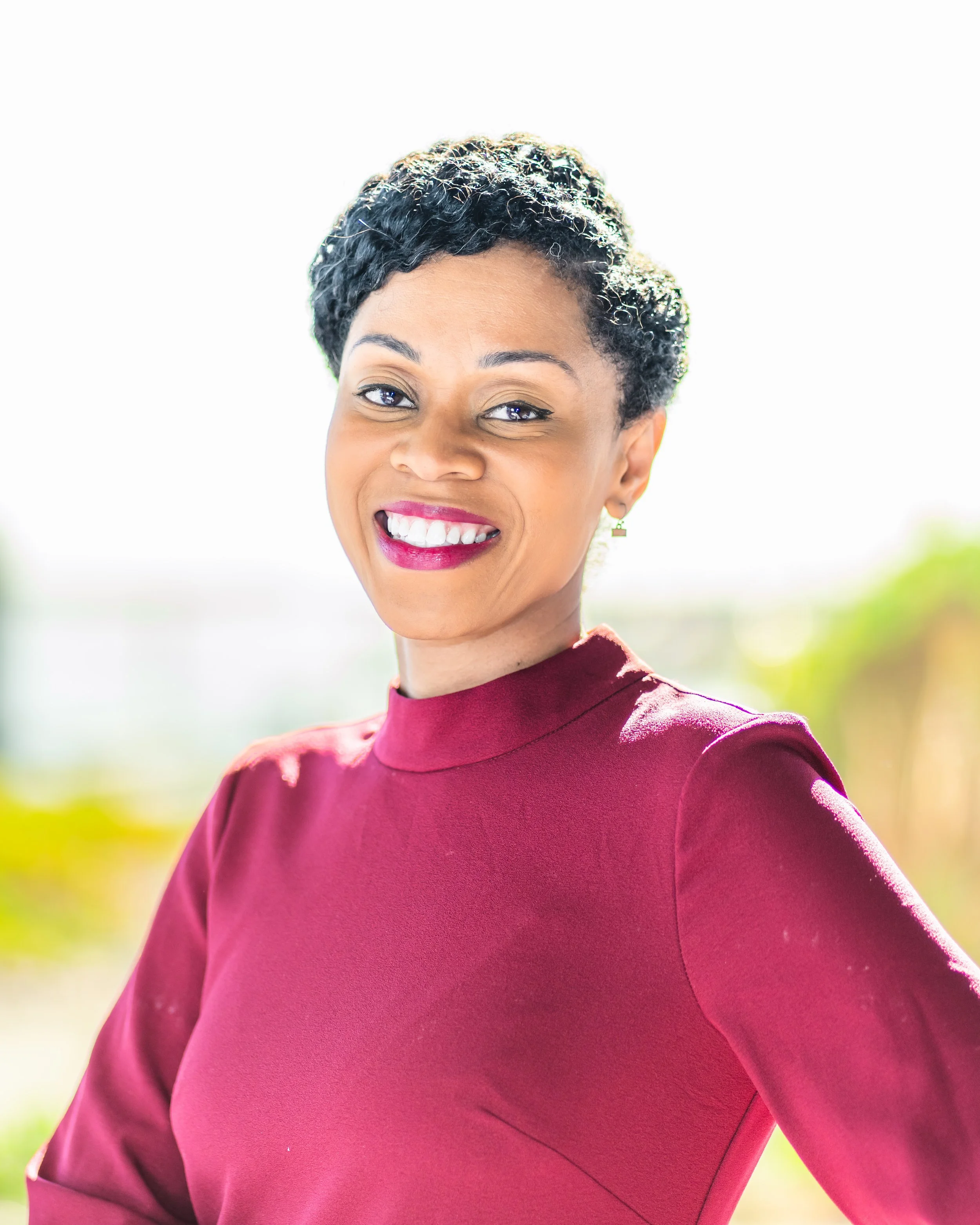 A smiling woman with short, curly hair wearing a burgundy top outdoors on a sunny day.