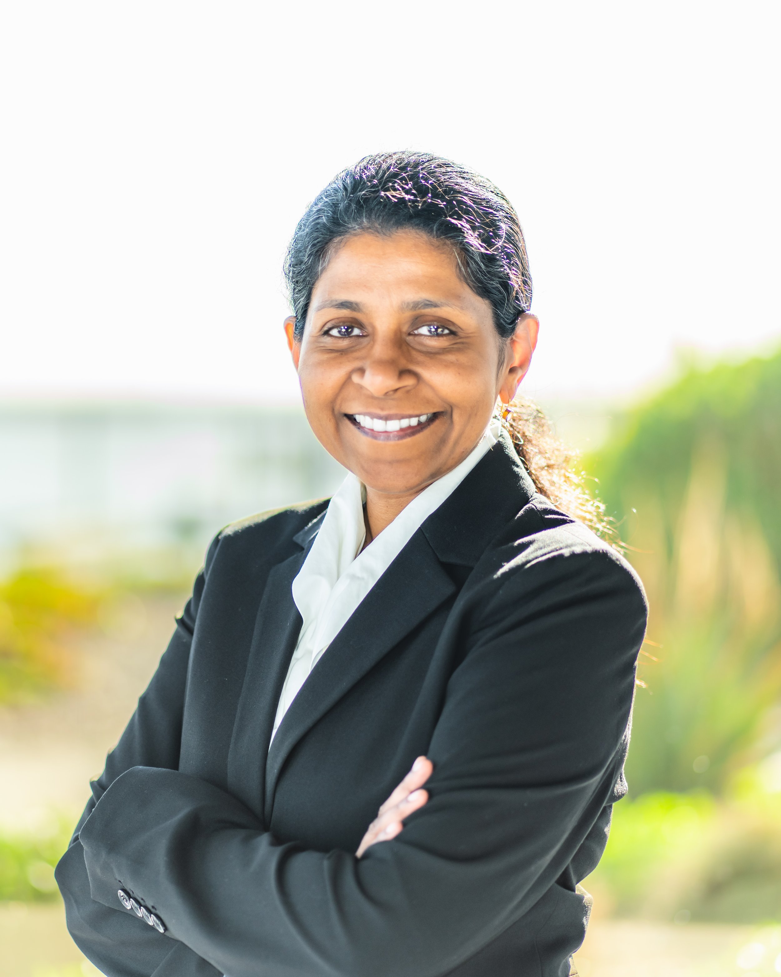 A smiling woman in a black suit and white blouse standing outdoors with arms crossed, with a blurred green and yellow natural background.