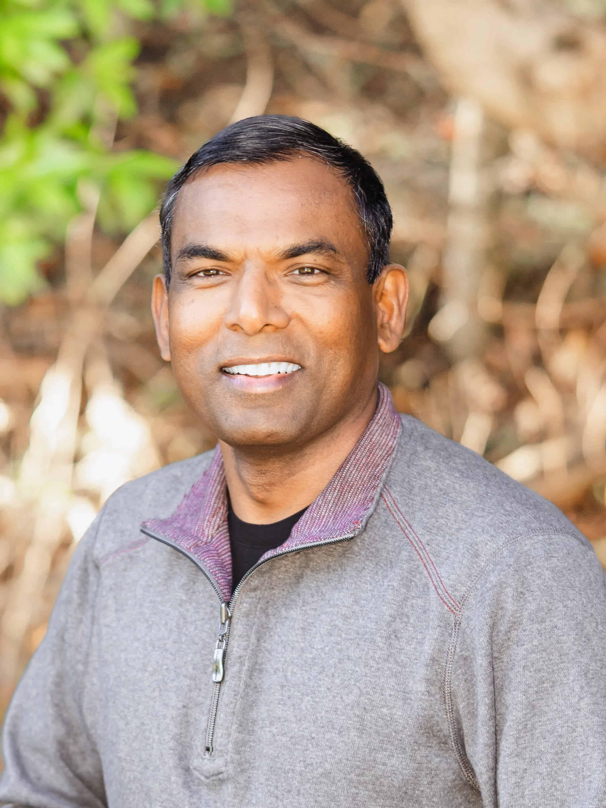 A man with short black hair, brown eyes, and medium skin tone smiling outdoors in front of a blurred natural background.