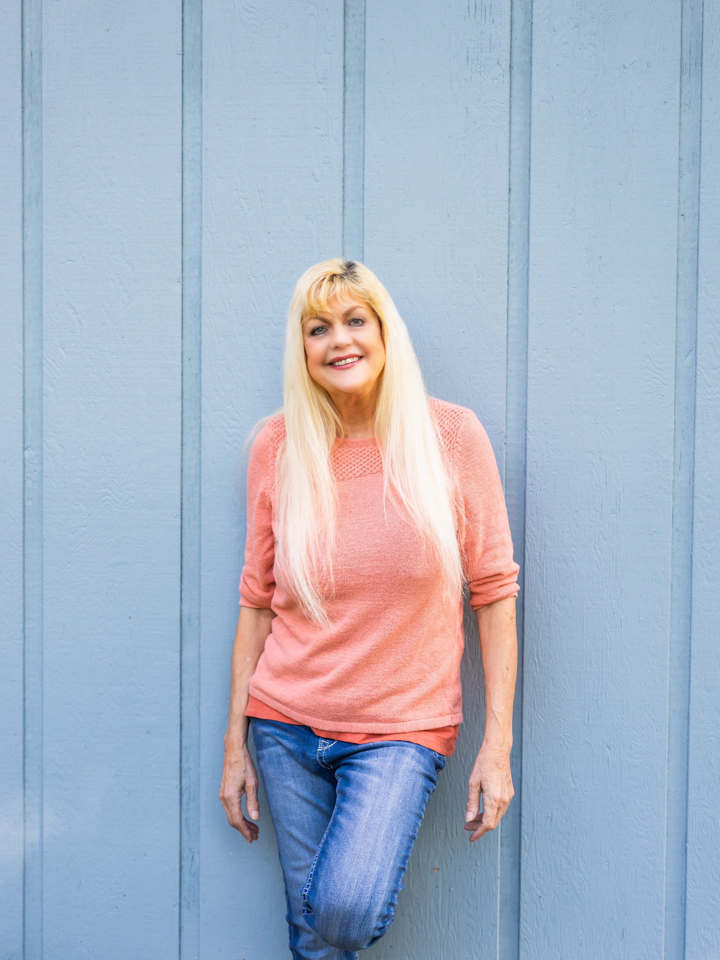 A woman with long blonde hair and a pink sweater smiling while standing against a light blue wooden wall.