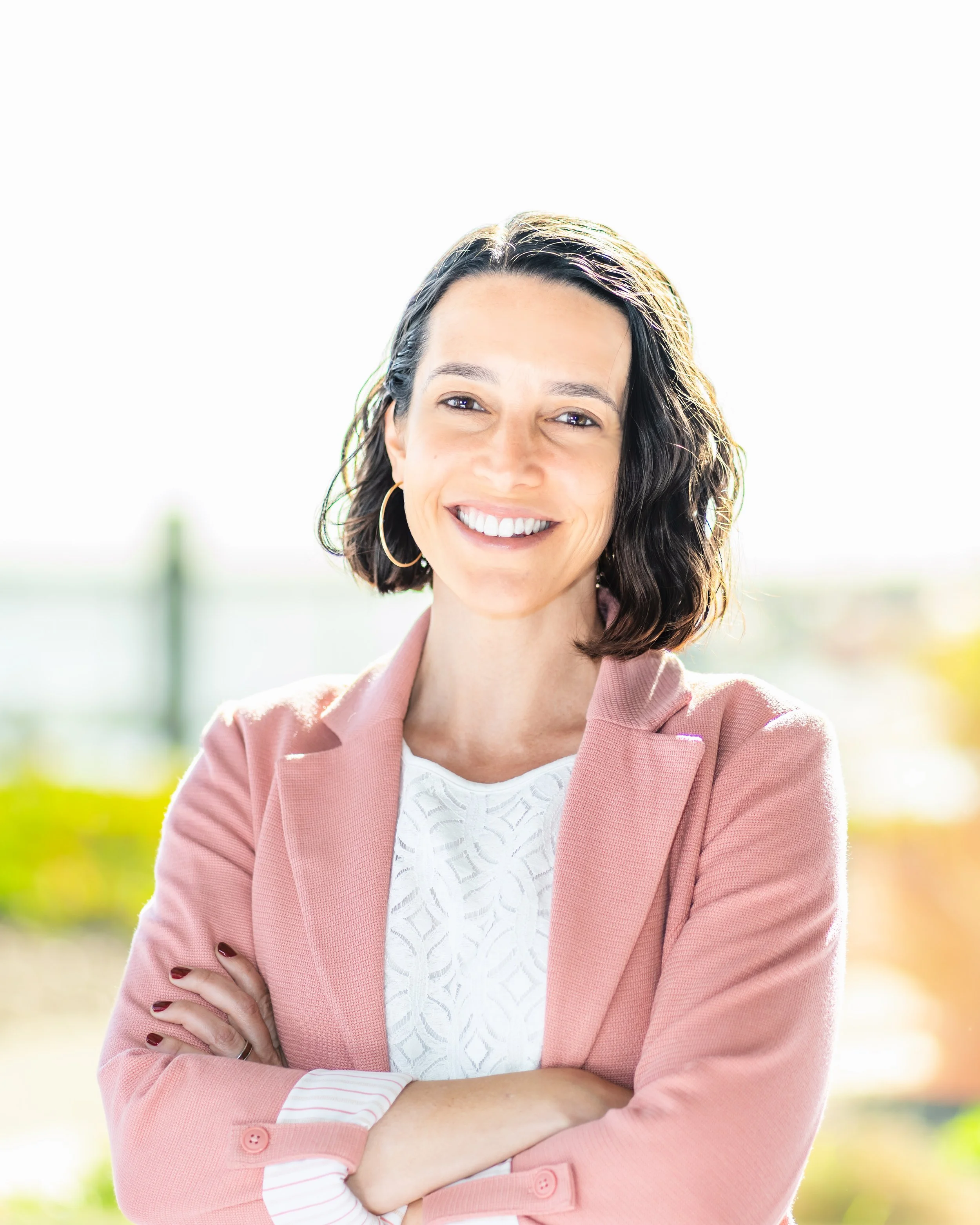 Smiling woman with dark, wavy hair, wearing a pink blazer and white top, standing outdoors with crossed arms.