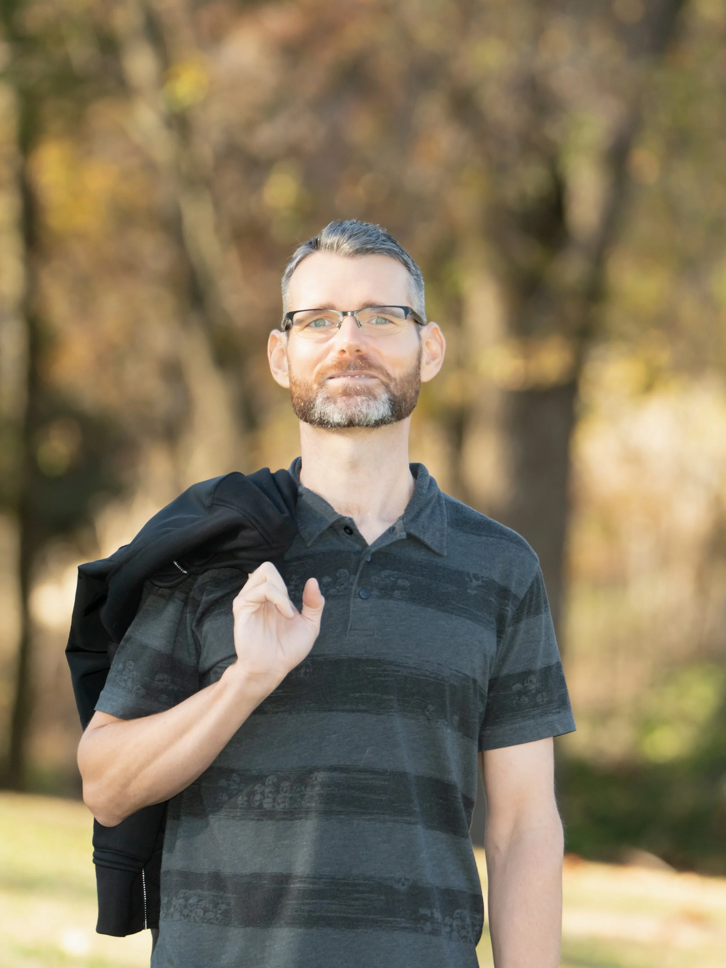 A man wearing glasses and a dark striped polo shirt carries a black jacket over his shoulder while standing outdoors with trees in the background.