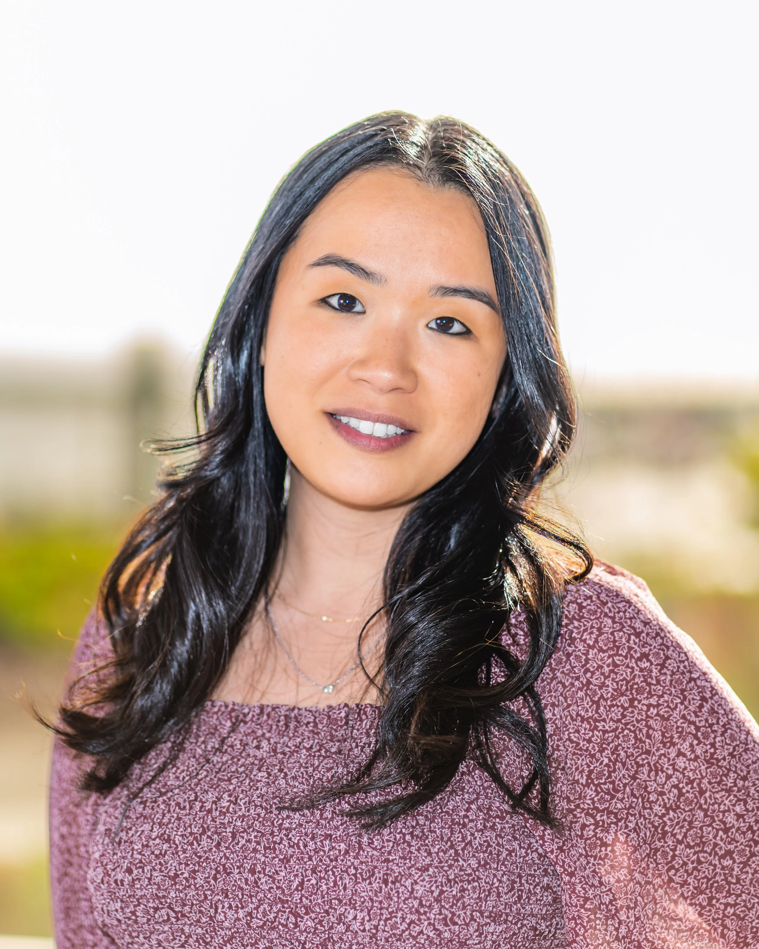 A woman with long black hair, wearing a maroon patterned top and a delicate necklace, smiling outdoors.