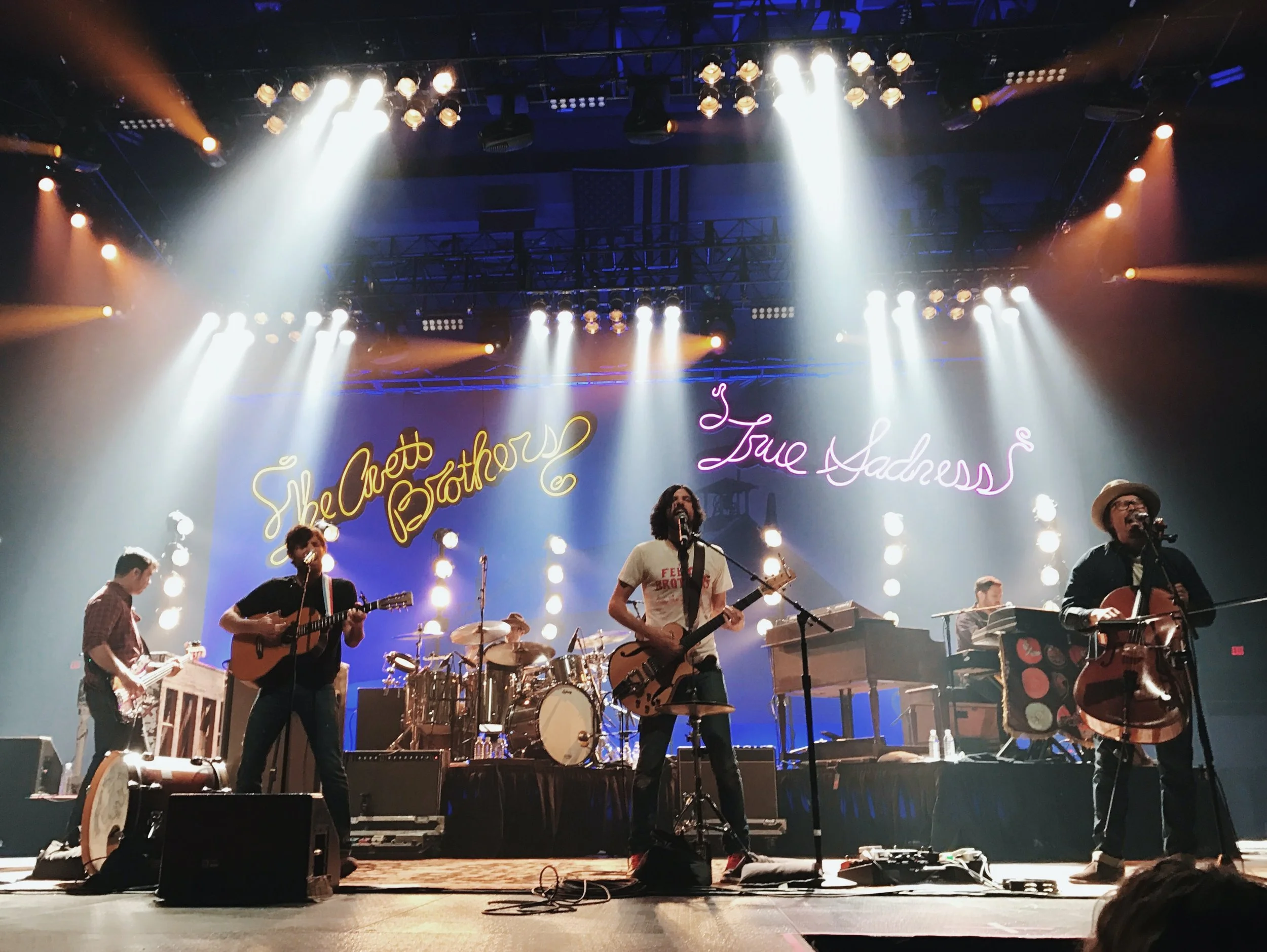 Band performing on stage with neon signs backdrops reading 'She Doesn't Like the Rain' and 'True Sadness', with multiple performers playing guitars and drums under stage lights.