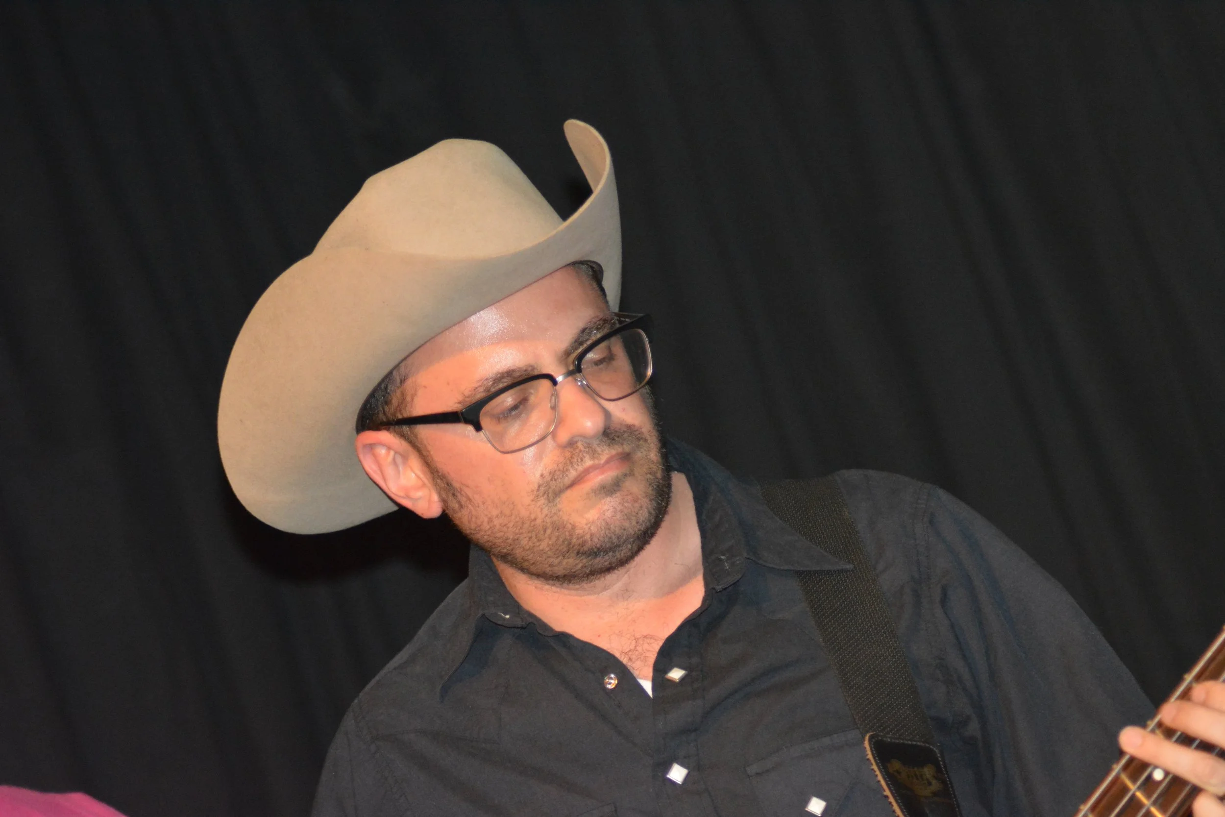A man wearing a large beige cowboy hat, glasses, and a dark shirt, appears to be playing a guitar against a black background.