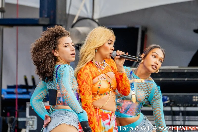 Three female performers on stage, one holding a microphone, wearing colorful, revealing outfits, with a background of stage equipment and outdoor setting.