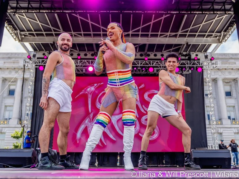 Three performers on stage wearing colorful, mesh, rainbow-themed outfits at an outdoor event, with a bright pink backdrop and stage lighting.