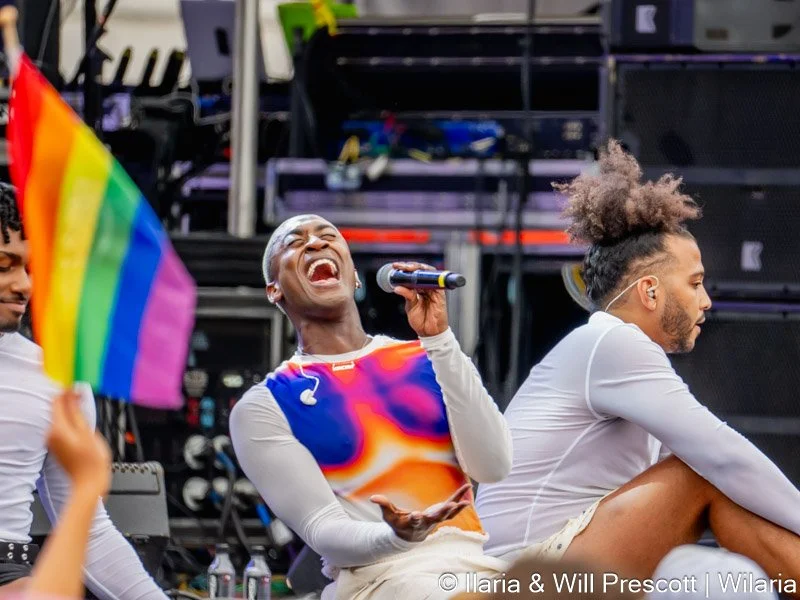 Two people sitting on stage at a pride event, one holding a rainbow pride flag and the other singing into a microphone, with colorful stage equipment in the background.