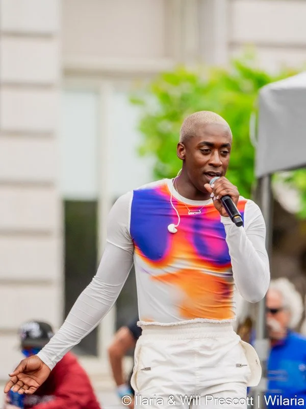 A young man with platinum blonde hair singing into a microphone during an outdoor event, wearing a colorful athletic top and white pants.