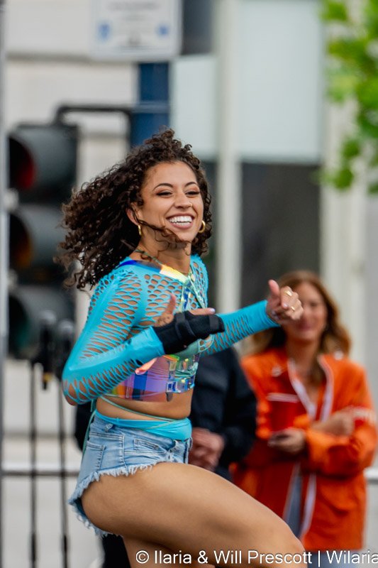 A smiling woman in a vibrant, colorful crop top and denim shorts, dancing outdoors with others in the background.